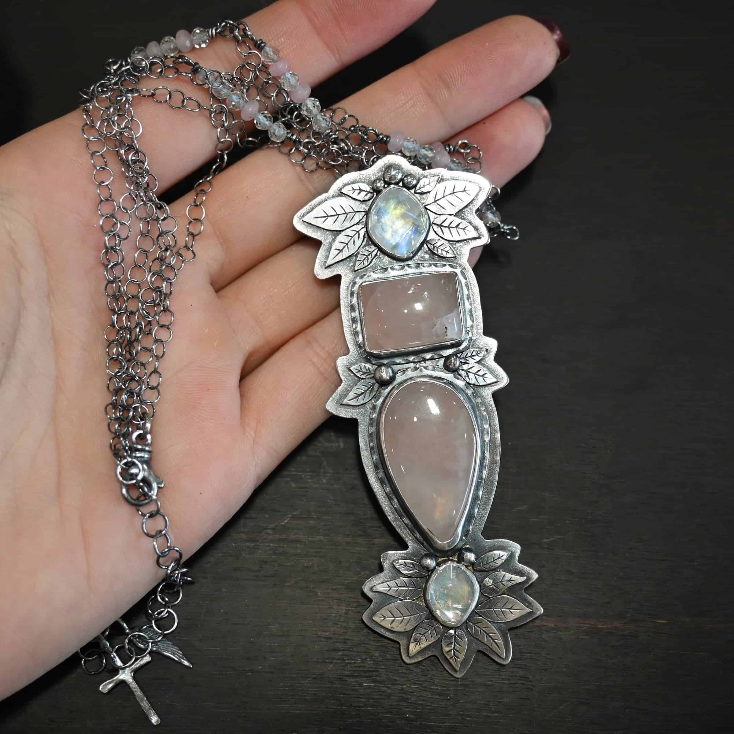 The Sparkling Lady Necklace Close up of the "Sparkling Lady" Necklace. Made out of sterling silver, rose quartz, and moonstone. There are two rose quartz and two moonstone. The top of the pendant has an oval moonstone, with a square rose quartz beneath it. Beneath that is another rose quartz that is a teardrop shape with the point facing downwards. At the bottom of the pendant is another moonstone that is an oval shape. Surrounding each of these stones and around the entire pendant are silver leaves and dots. Pendant is laying against the fingertips of a hand.