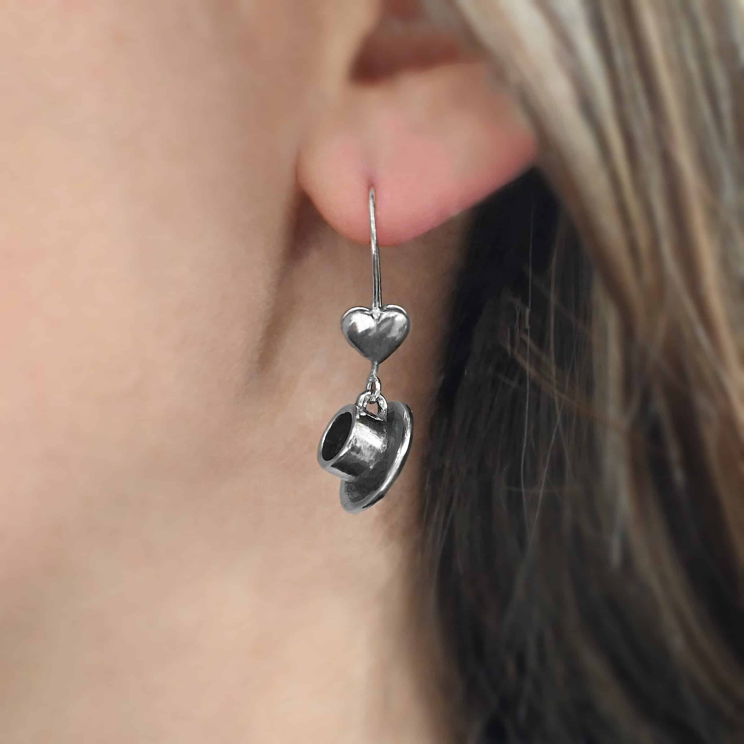 Photo of the "True Addiction" Coffee Earring as shown up close by a model. Made out of sterling silver. Earring has an oxidized french hook ear wire. The bottom of each earwire has a sterling silver heart attached to it. Attached to the bottom of the heart is a small semicircle loop. The handle of a cup is attached to it, along with a small saucer.