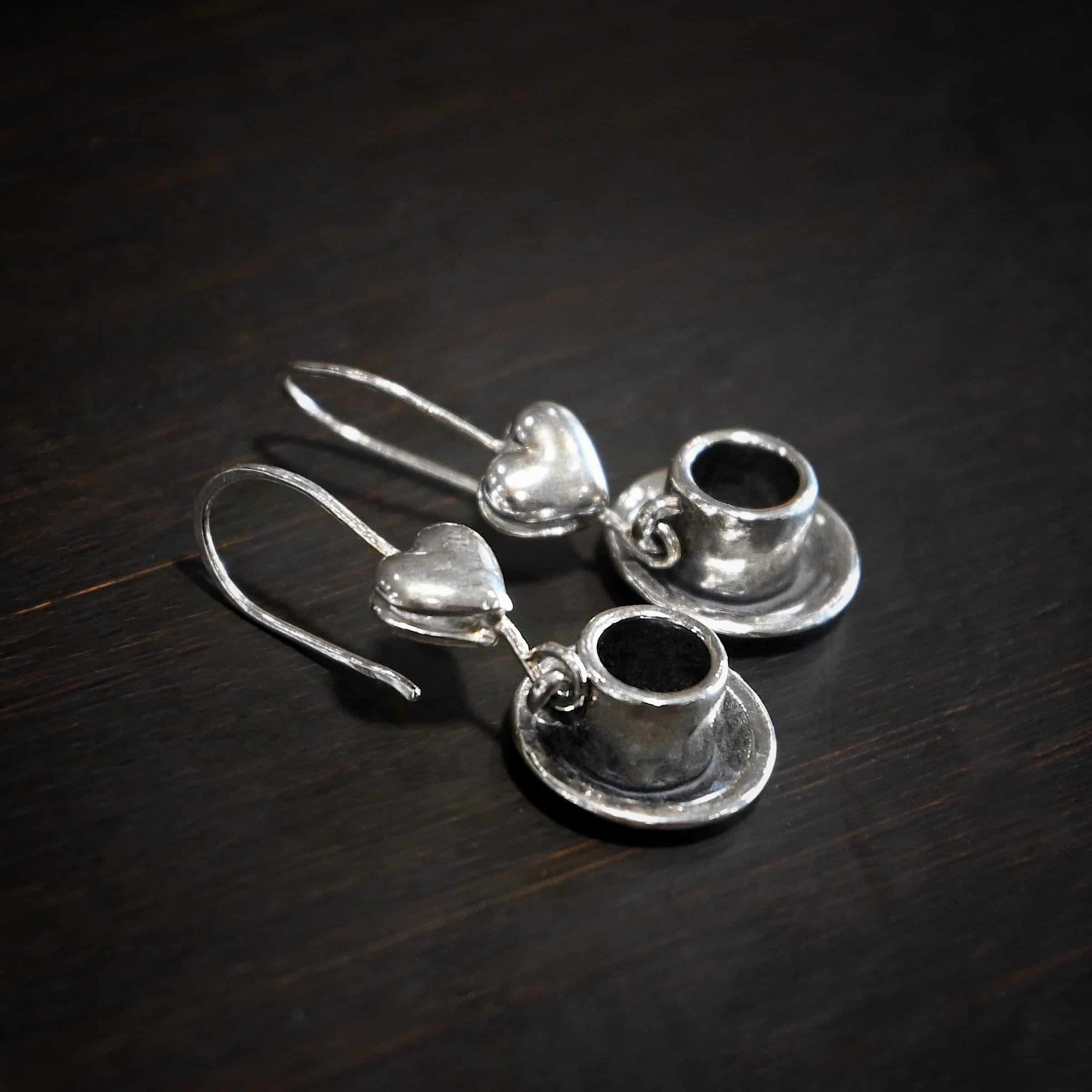 Photo of the "True Addiction" Coffee Earrings. Made out of sterling silver. Earrings have oxidized french hook ear wires. The bottom of each earwire has a sterling silver heart attached to it. Attached to the bottom of the heart is a large semicircle loop. In the center of the loop, attached on each side is a saucer and a coffee cup. Earrings are lying against a dark background.