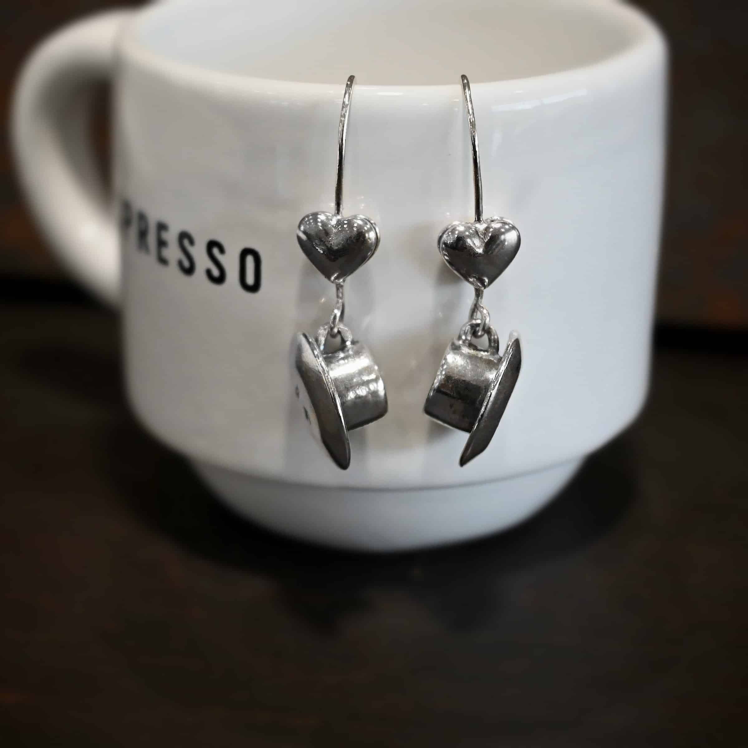 Photo of the "True Addiction" Coffee Earrings. Made out of sterling silver. Earrings have oxidized french hook ear wires. The bottom of each earwire has a sterling silver heart attached to it. Attached to the bottom of the heart is a large semicircle loop. In the center of the loop, attached on each side is a saucer and a coffee cup. Earrings are hanging off of a small espresso cup.