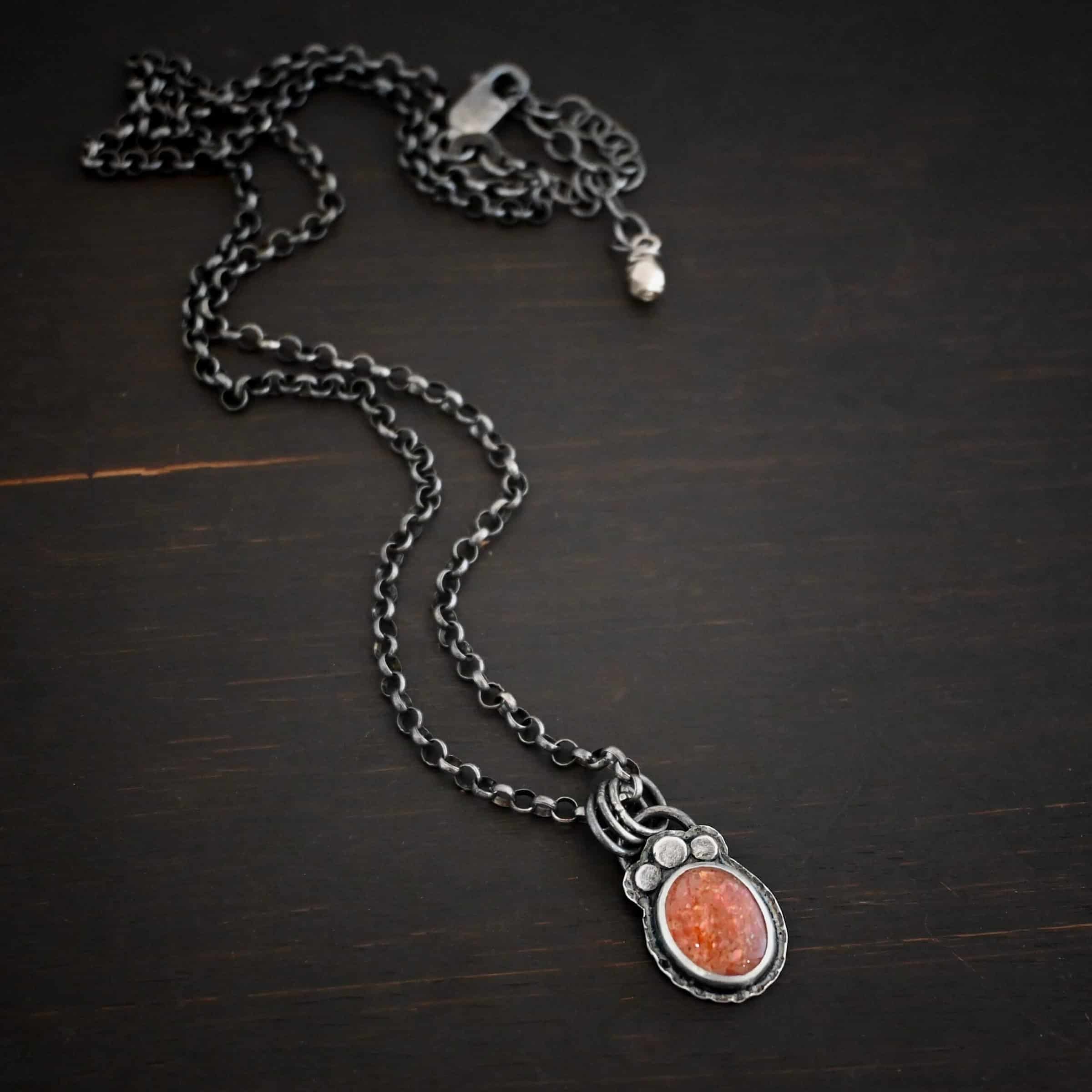 Photo of the "Ileah" Necklace. There is a small oval sunstone in the center of the pendant. There are three hammered silver dots on the top of it that follow the curve of the top of the oval. There is a loop soldered to the back with three silver jump rings going through it. The chain of the necklace goes through the jump rings. Necklace is lying against a dark background.