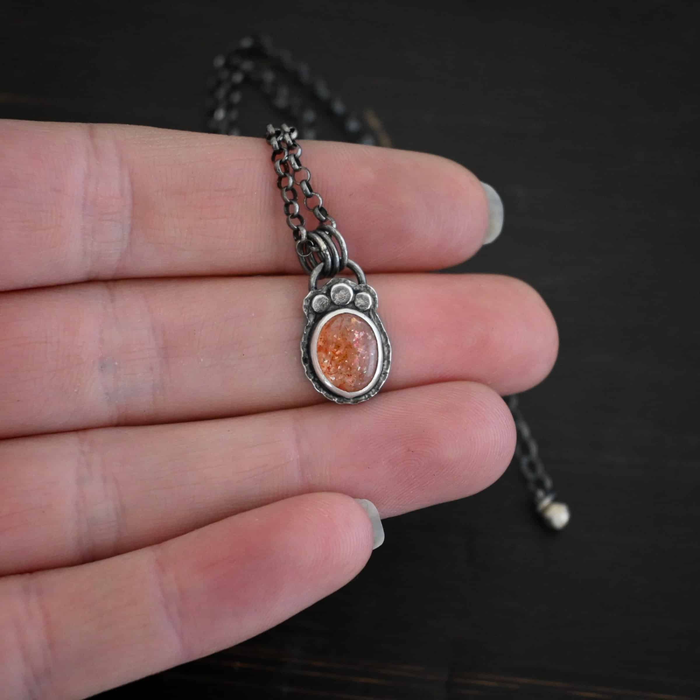 Photo of the "Ileah" Necklace. There is a small oval sunstone in the center of the pendant. There are three hammered silver dots on the top of it that follow the curve of the top of the oval. There is a loop soldered to the back with three silver jump rings going through it. The chain of the necklace goes through the jump rings. Necklace is lying against the fingertips of a hand.