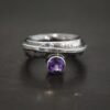 Photo of the "Mistletoe" Ring. Made out of sterling silver and amethyst. There is a small circular amethyst attached to the bottom of a ring band. It is a deep purple color. The ring band is thick and hammered. There is a thick line of silver that the top is hammered flat, but still sticks up from the band and that is soldered and wrapped around the band. Ring is sitting against a dark background.