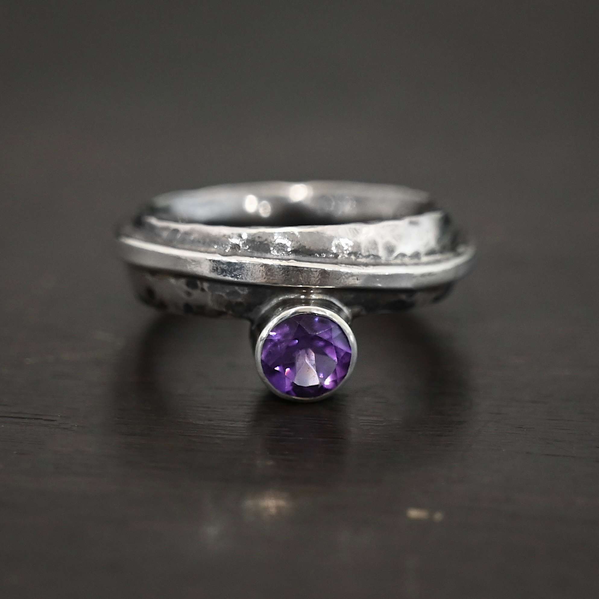 Photo of the "Mistletoe" Ring. Made out of sterling silver and amethyst. There is a small circular amethyst attached to the bottom of a ring band. It is a deep purple color. The ring band is thick and hammered. There is a thick line of silver that the top is hammered flat, but still sticks up from the band and that is soldered and wrapped around the band. Ring is sitting against a dark background.