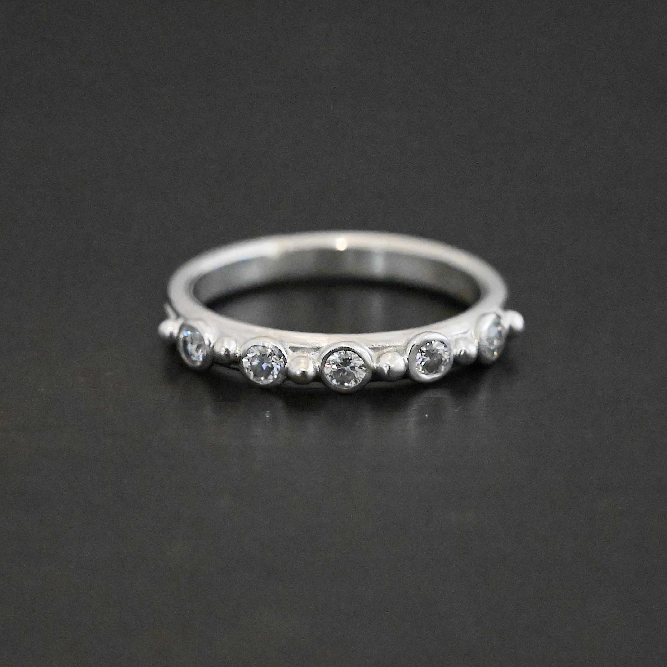 Close up of the "Years Of Love" Wedding Band. Made out of sterling silver and moissanites. There are five moissanite stones, each separated by a sterling silver dot. There are six sterling silver dots. One on each end of the moissanites to end the line, and four in between separating the other three moissanites. Ring is sitting against a dark background.