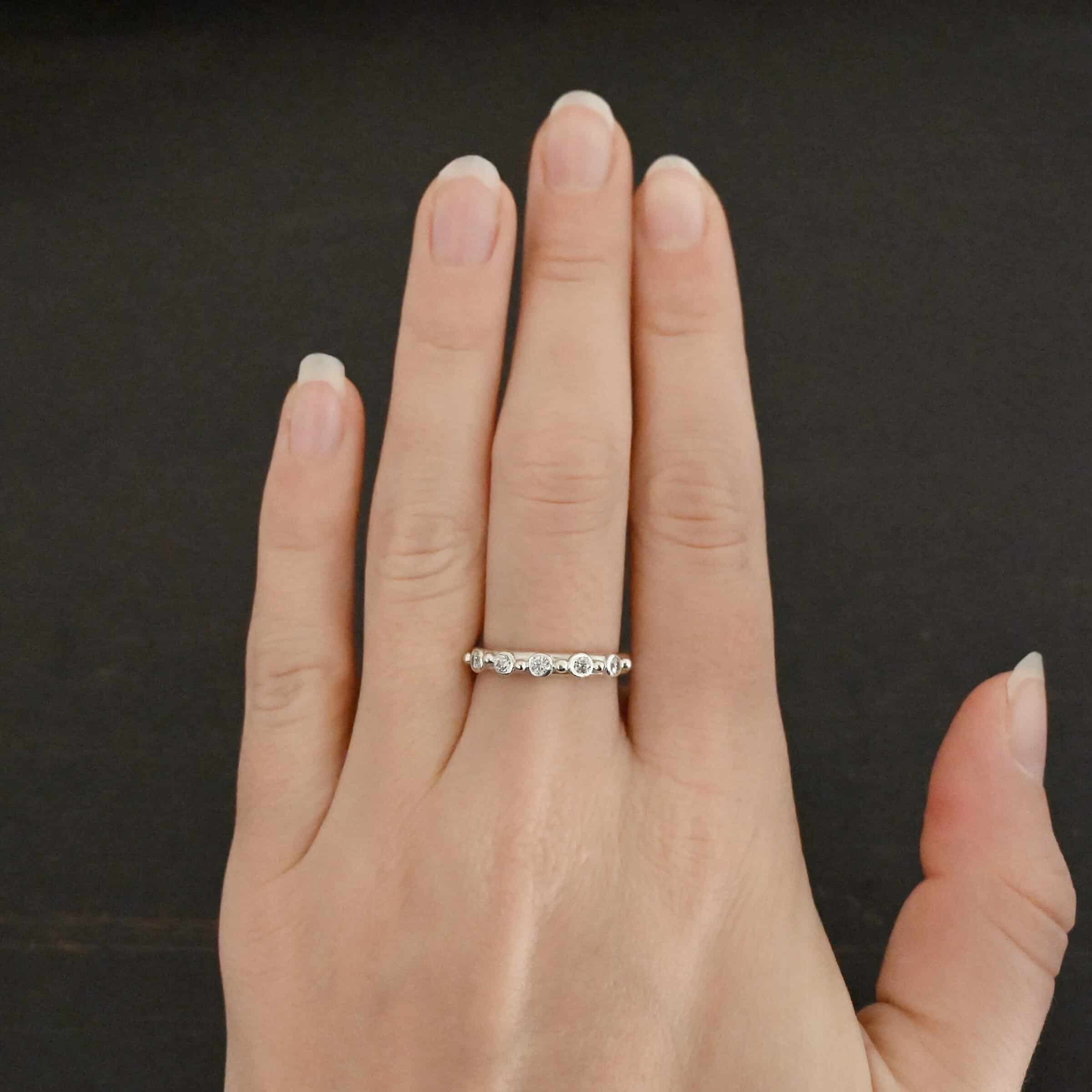 Close up of the "Years Of Love" Wedding Band. Made out of sterling silver and moissanites. There are five moissanite stones, each separated by a sterling silver dot. There are six sterling silver dots. One on each end of the moissanites to end the line, and four in between separating the other three moissanites. Ring is worn on a models middle finger to demonstrate size.