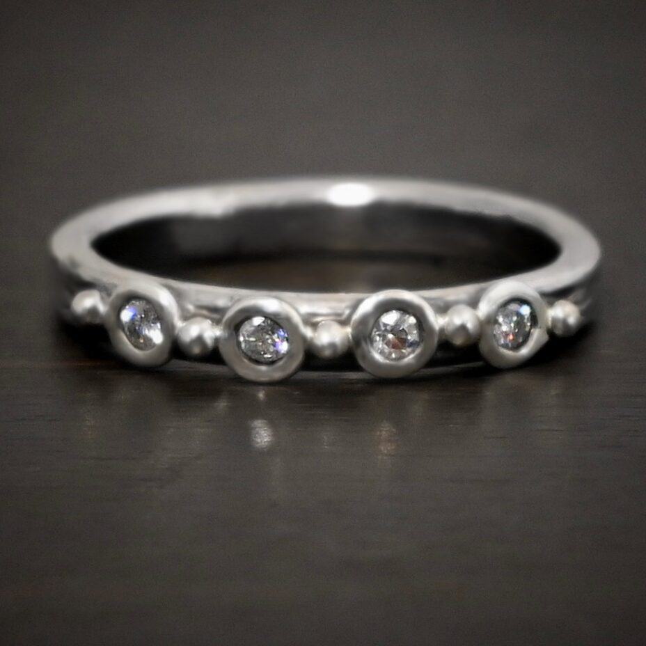 Photo of the "Serenity" Ring. The band is silver. There are four diamonds going across the center of the band. Each one is separated by a ball of silver. There are five balls of silver. Ring is sitting against a dark background.