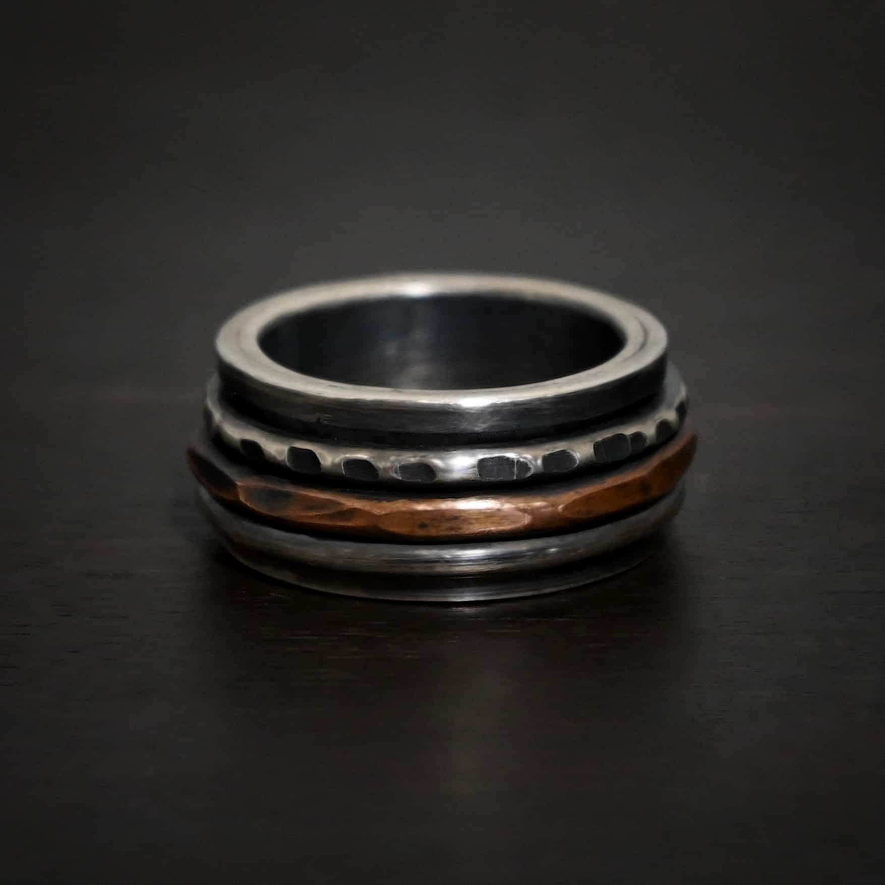 Revolver Ring Photo of the "Revolver" Ring. Made out of sterling silver, copper, and 14k gold. The top and bottom of the ring are silver, and there is a gap in the center of the ring. The gap is taken up by a hammered copper spinner, a thick, hammered sterling silver spinner, and a smooth silver spinner. Each band in the center spins around. Ring is sitting against a dark background.