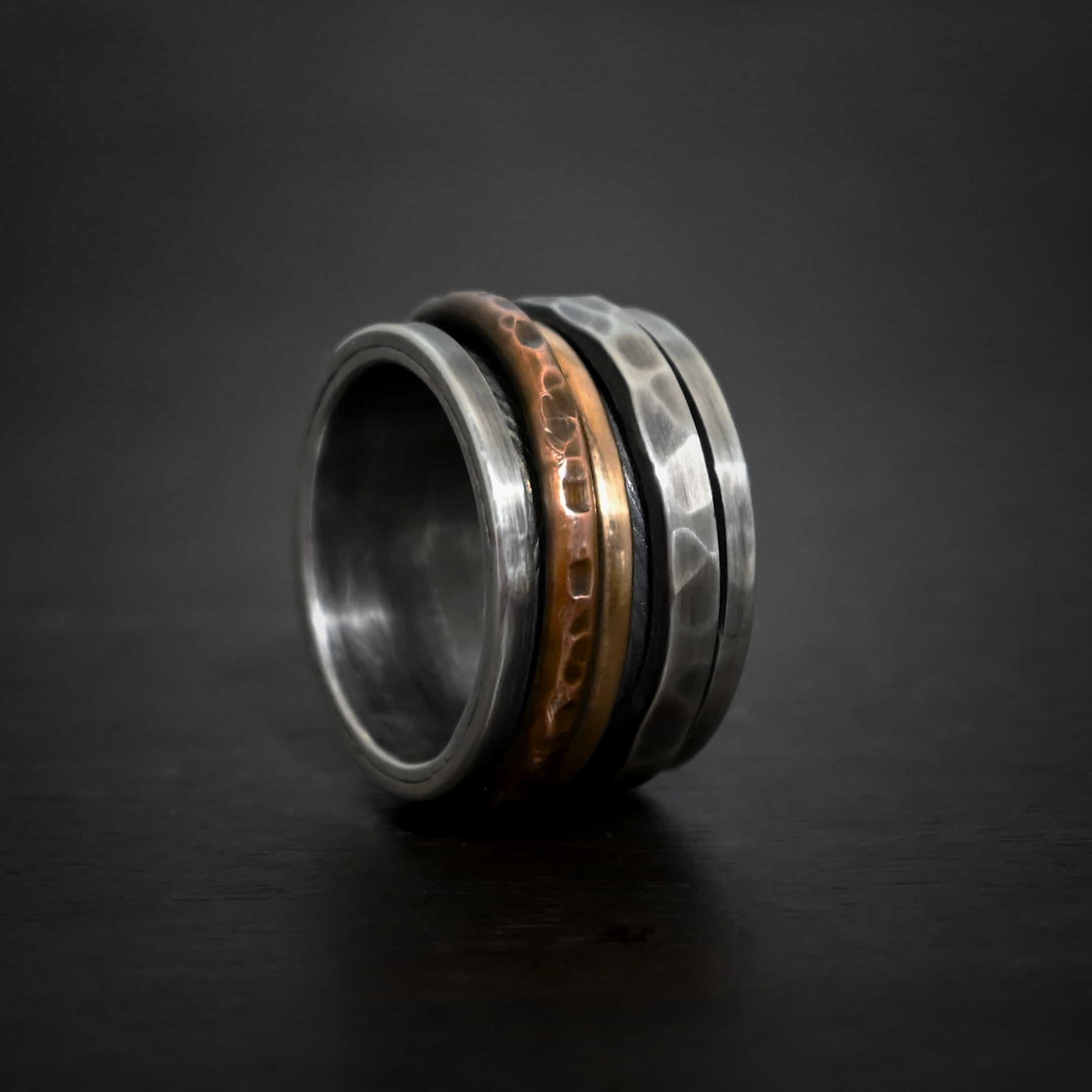 Royal Revolver Ring Photo of the "Royal Revolver" Ring. Made out of sterling silver, copper, and 14k gold. The top and bottom of the ring are silver, and there is a gap in the center of the ring. The gap is taken up by a hammered copper spinner, a smooth 14k gold spinner, and a thick, hammered sterling silver spinner. Each band in the center spins around. Ring is sitting against a dark background.