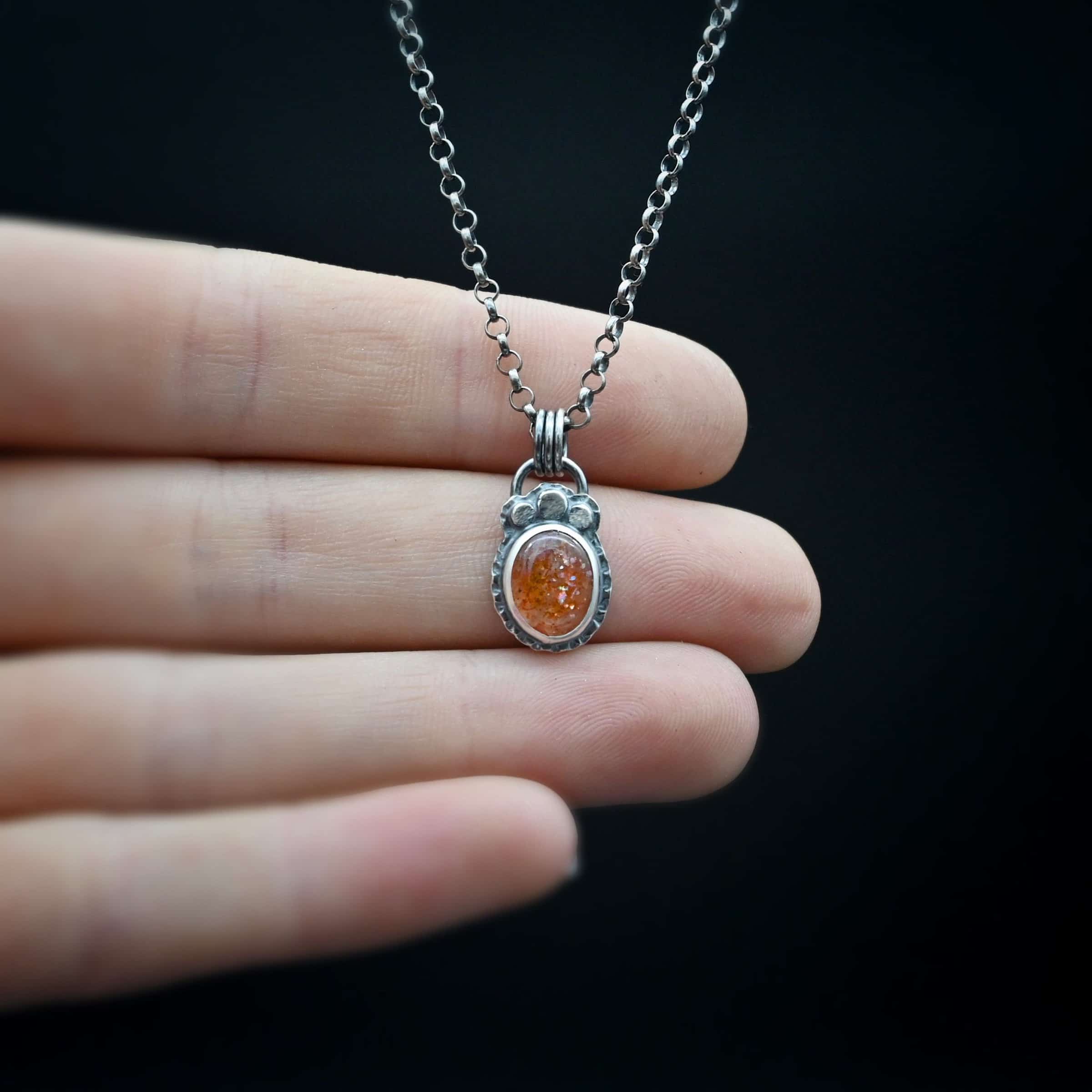 Photo of the "Ileah" Necklace. There is a small oval sunstone in the center of the pendant. There are three hammered silver dots on the top of it that follow the curve of the top of the oval. There is a loop soldered to the back with three silver jump rings going through it. The chain of the necklace goes through the jump rings. Necklace is lying against the fingertips of a hand.
