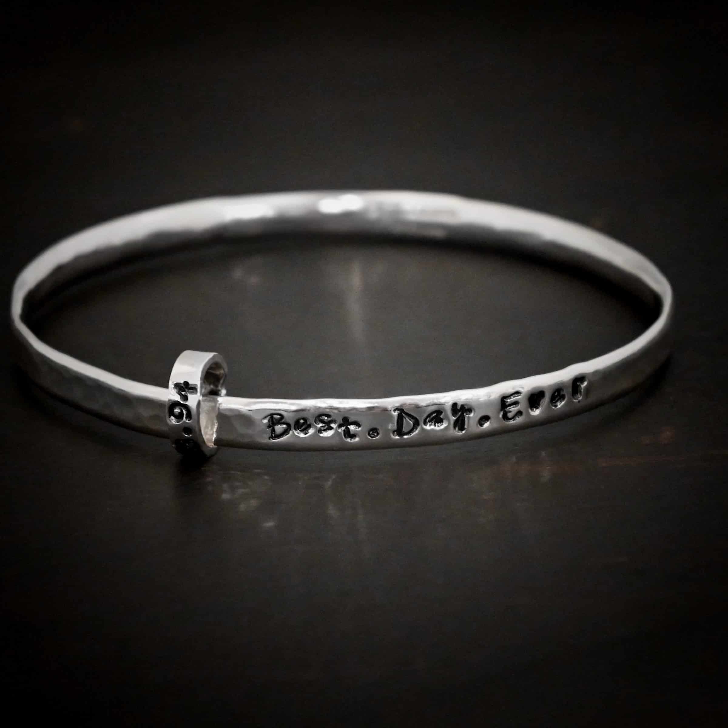 Best Day Ever Bangle Photo of the "Best Day Ever" Bangle. Made out of sterling silver. The bangle is hammered and shiny. There is one thick jump ring on the left side of it with an oxidized date hammered into it. The words "Best. Day. Ever" are also oxidized and hammered across the front of the bangle. Bangle is sitting against a dark background.