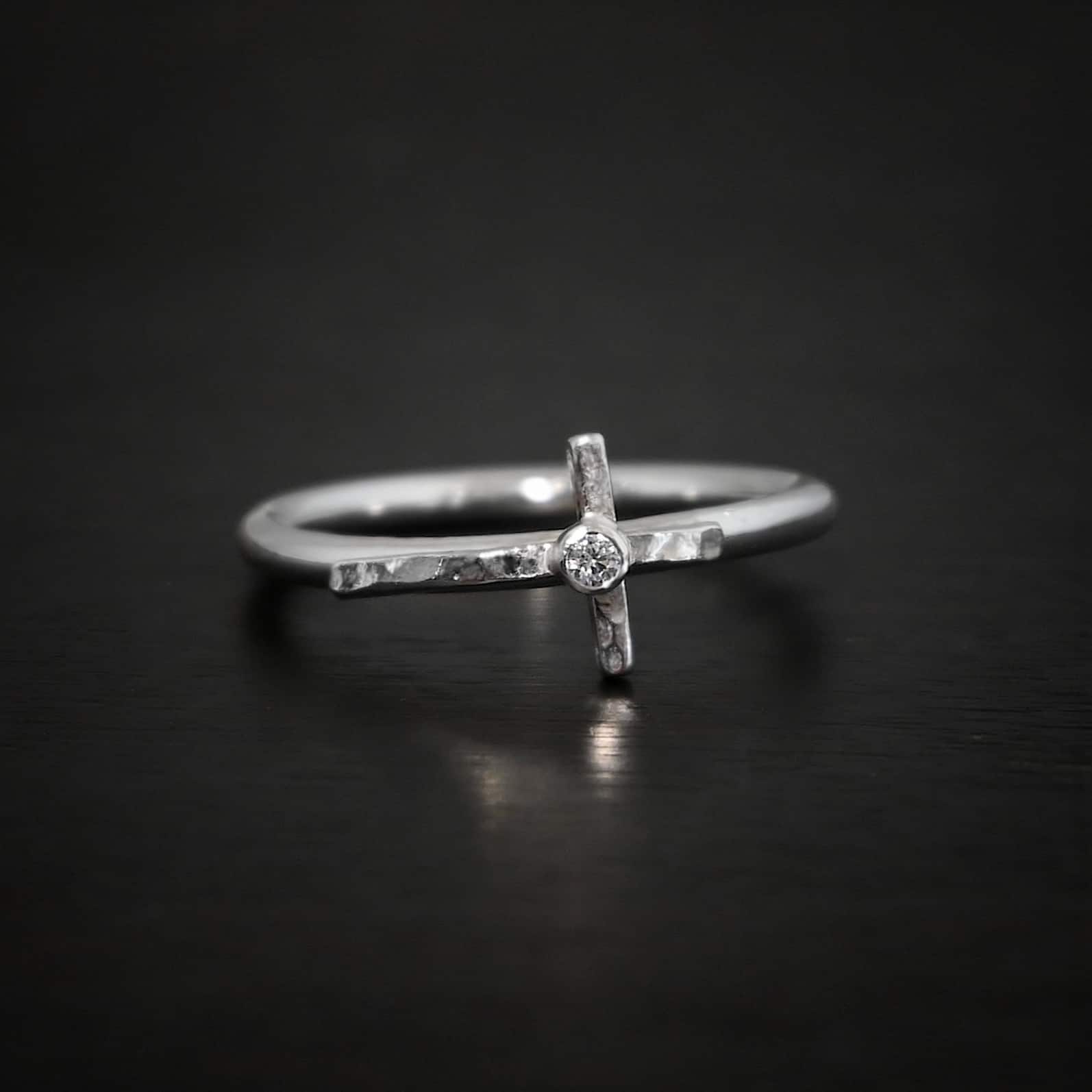 Photo of a creator ring. Ring is made out of sterling silver. The ring band is a thin circle. There is a cross that is in the center of the ring band and set horizontally. The top of bottom of the cross are soldered onto the ring band. The top side of the cross is sticking into the air and the bottom side of the cross is set on the table. There is a small cubic zirconia in the center of the cross. Ring is sitting against a dark background.