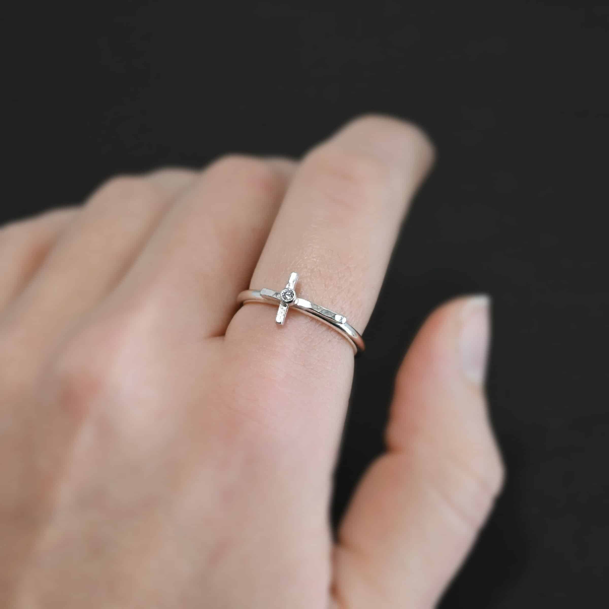 Photo of a creator ring. Ring is made out of sterling silver. The ring band is a thin circle. There is a cross that is in the center of the ring band and set horizontally. The top of bottom of the cross are soldered onto the ring band. The top side of the cross is sticking into the air and the bottom side of the cross is set on the table. There is a small cubic zirconia in the center of the cross. Ring is sitting worn by a model up close.