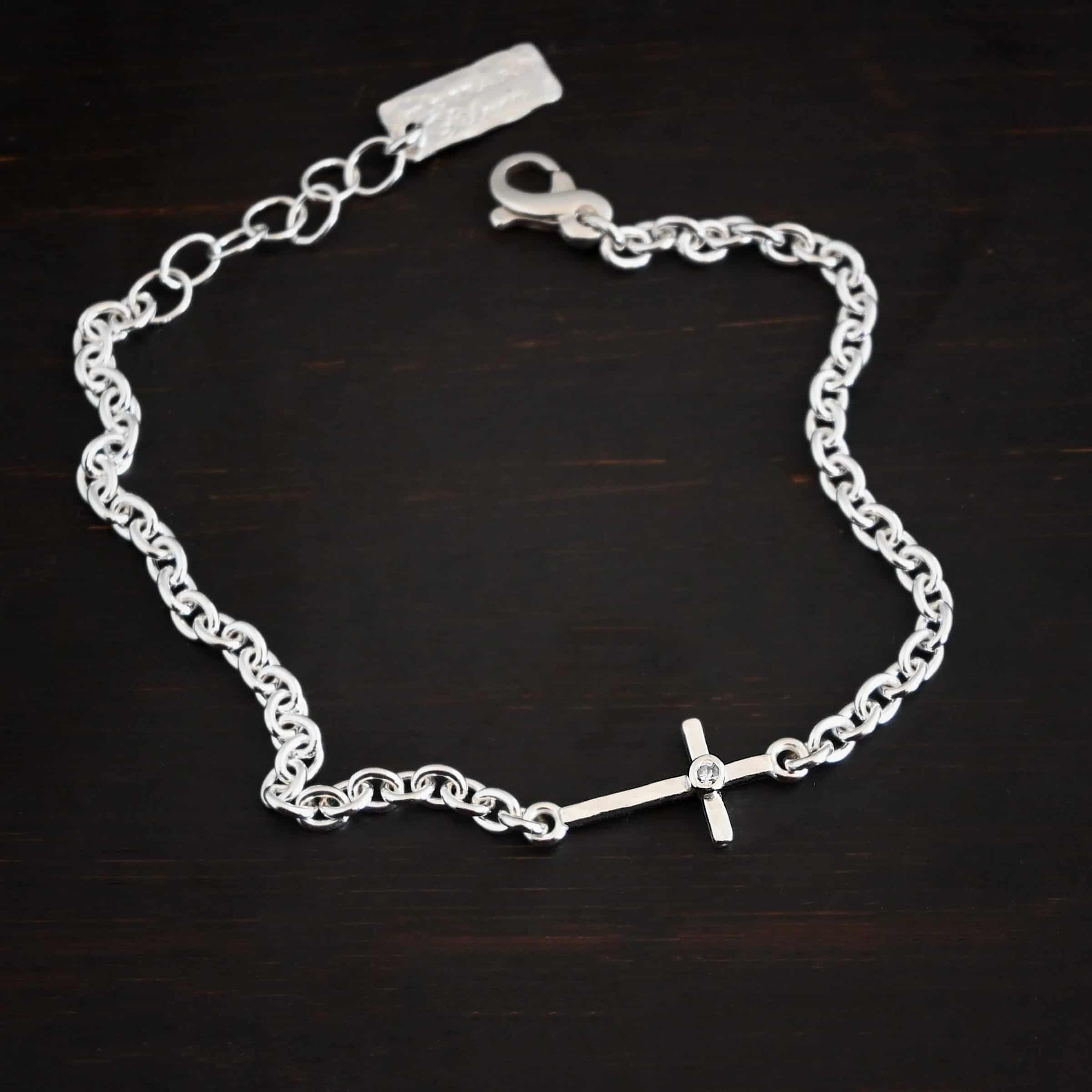 Photo of the "Creator" Bracelet. There is a cross set horizontally with a loop on each end. Each end of the cross is connected to each side of the chain in the center of the bracelet. There is a small cz in the center of the cross. The bracelet is shiny, and lying against a dark background.