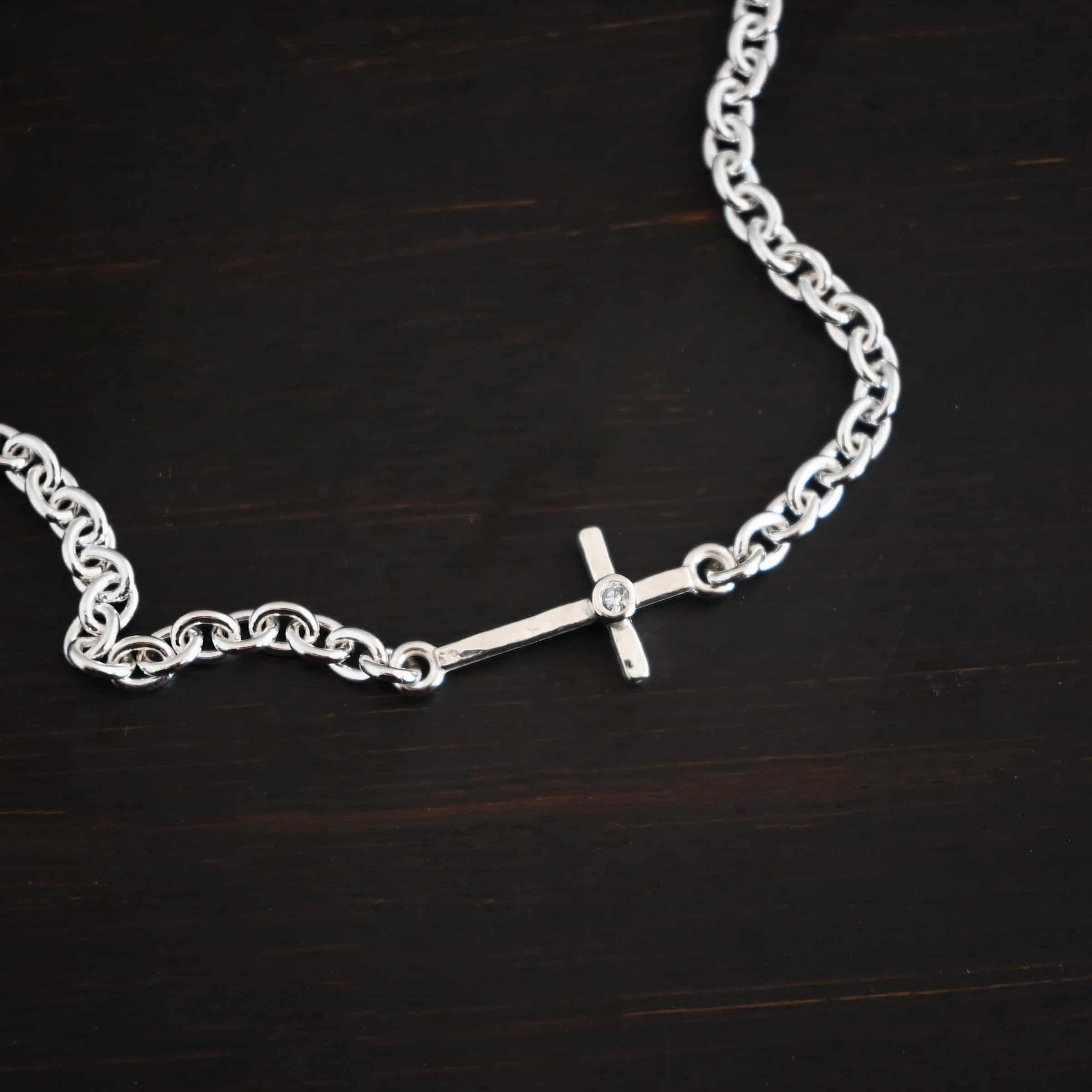 Photo of the "Creator" Bracelet. There is a cross set horizontally with a loop on each end. Each end of the cross is connected to each side of the chain in the center of the bracelet. There is a small cz in the center of the cross. The bracelet is shiny, and lying against a dark background.