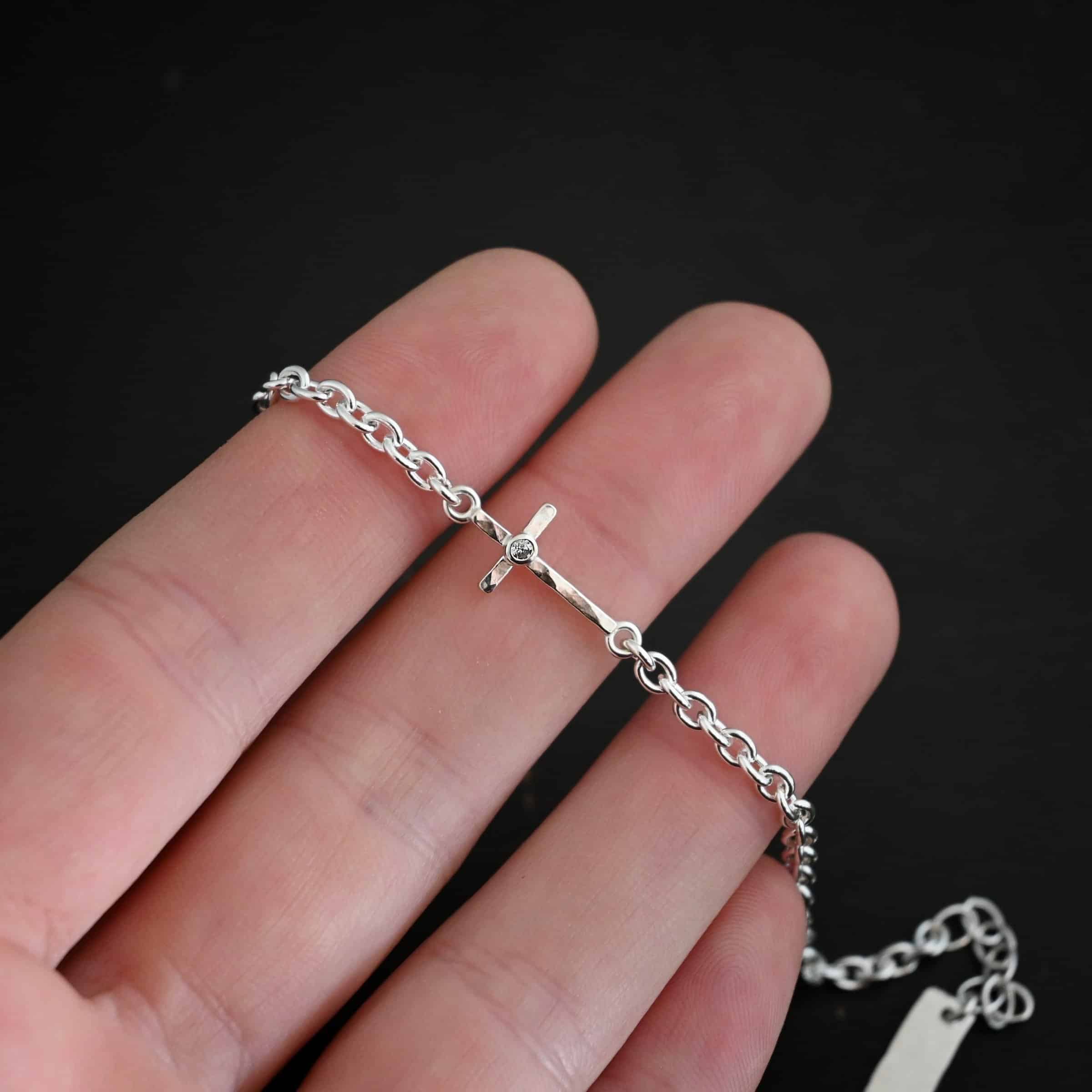 Photo of the "Creator" Bracelet. There is a cross set horizontally with a loop on each end. Each end of the cross is connected to each side of the chain in the center of the bracelet. There is a small cz in the center of the cross. The bracelet is shiny, and lying against the fingertips of a hand.