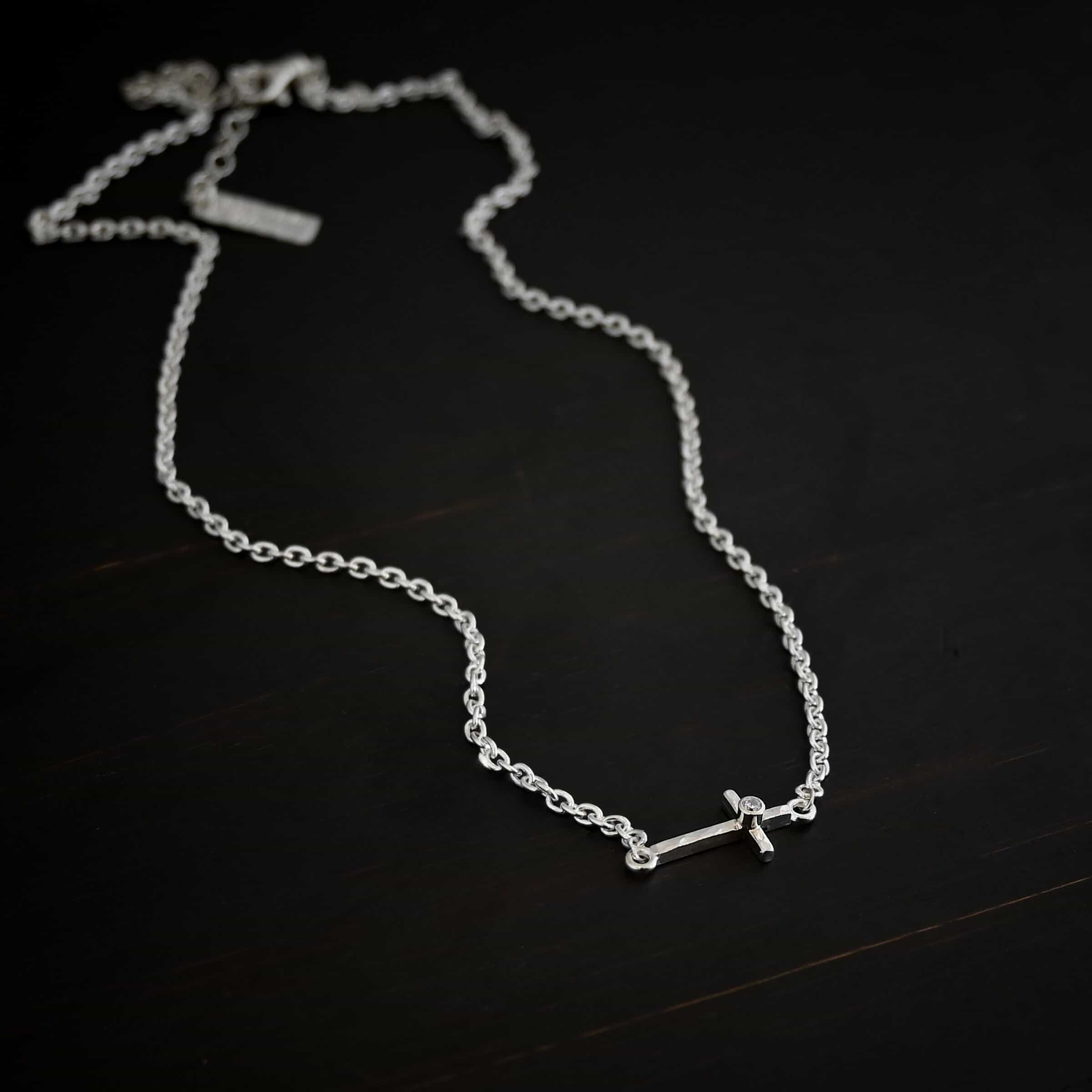 Creator Necklace shiny cz Photo of the "Creator" Necklace. Made out of sterling silver. There is a small, but thick hammered silver cross hung vertically and attached on both ends to the chain of the necklace. There is a small cz in the center of the cross. Each end of the cross has a loop on it to attach each end to the chain. Necklace is shiny and lying against a dark background.