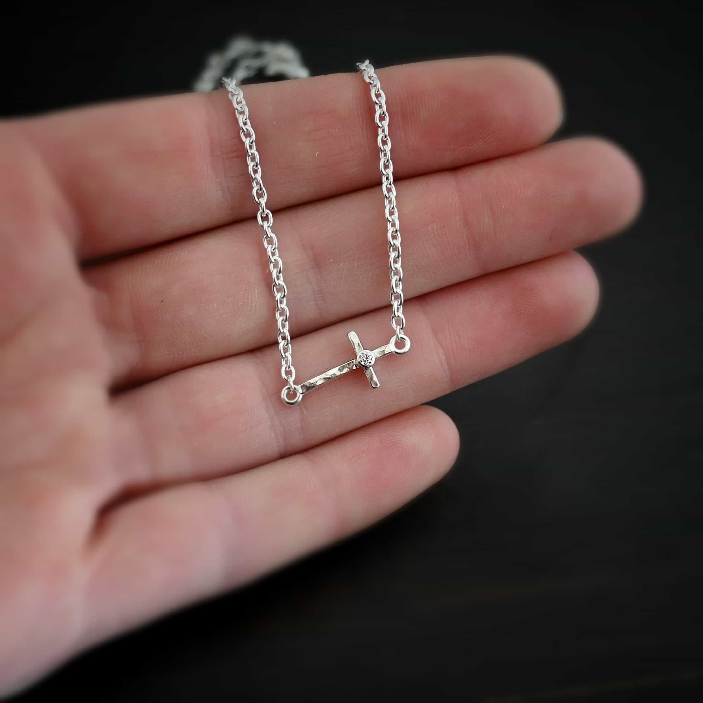 Creator Necklace shiny cz Photo of the "Creator" Necklace. Made out of sterling silver. There is a small, but thick hammered silver cross hung vertically and attached on both ends to the chain of the necklace. There is a small cz in the center of the cross. Each end of the cross has a loop on it to attach each end to the chain. Necklace is shiny and lying against the fingertips of a hand.