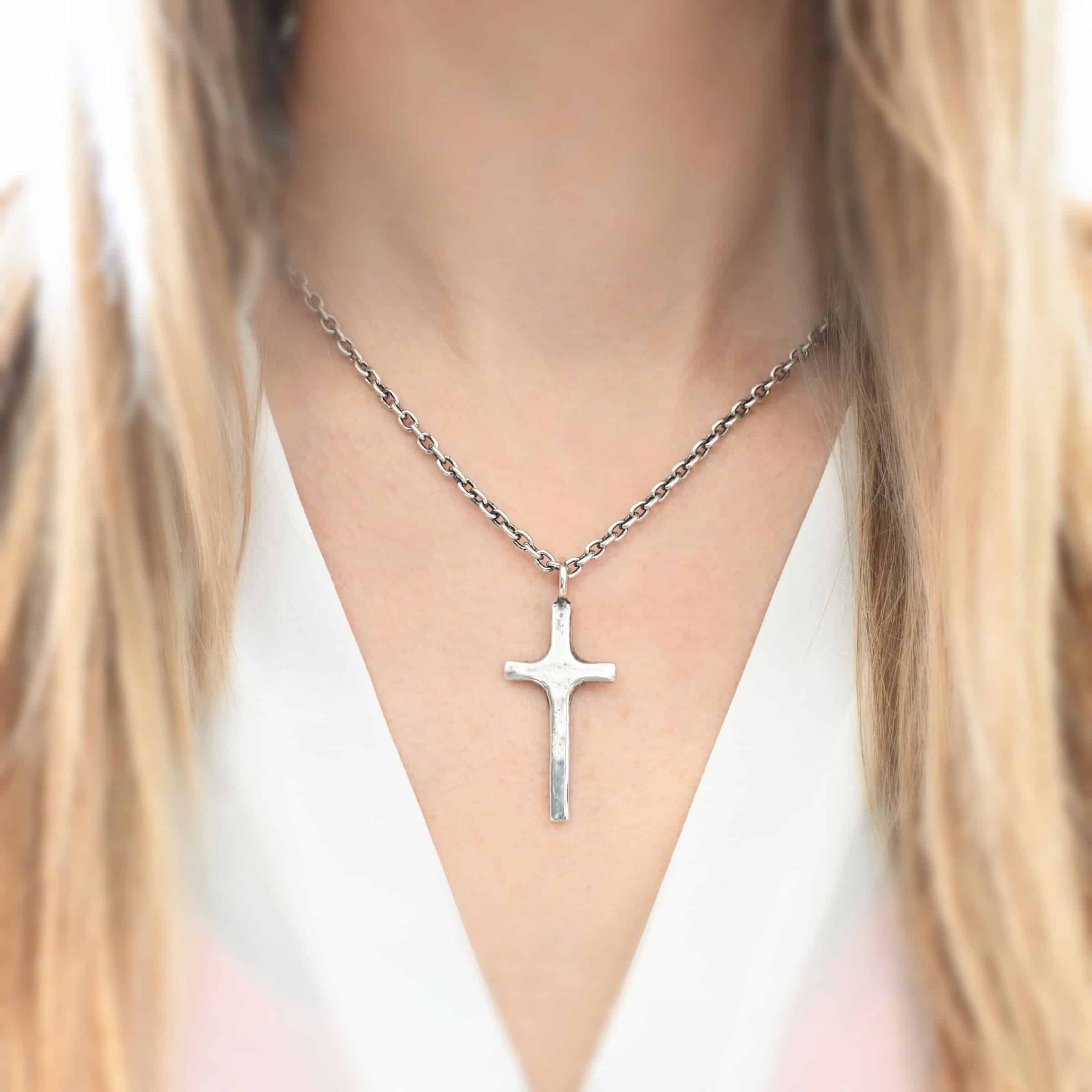 Photo of the "King Of Kings" Necklace. Made out of sterling silver. There is a medium sized cross that is made out of smooth, thick sterling silver. It is shiny and brushed lightly and has a loop soldered to the top of it. There is a chain that goes through the top of the necklace. Necklace is worn by a model. Necklace is shiny.