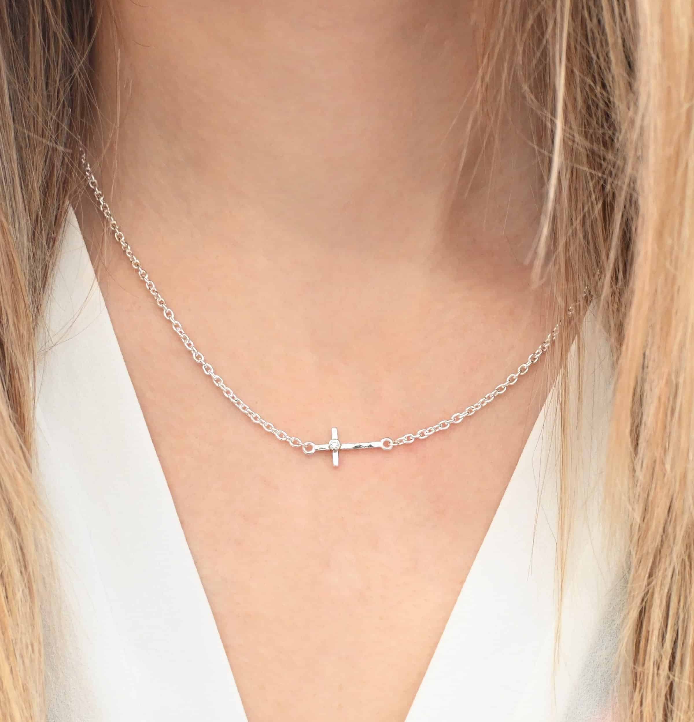 Creator Necklace shiny cz Photo of the "Creator" Necklace. Made out of sterling silver. There is a small, but thick hammered silver cross hung vertically and attached on both ends to the chain of the necklace. There is a small cz in the center of the cross. Each end of the cross has a loop on it to attach each end to the chain. Necklace is shiny and worn up close by a model.