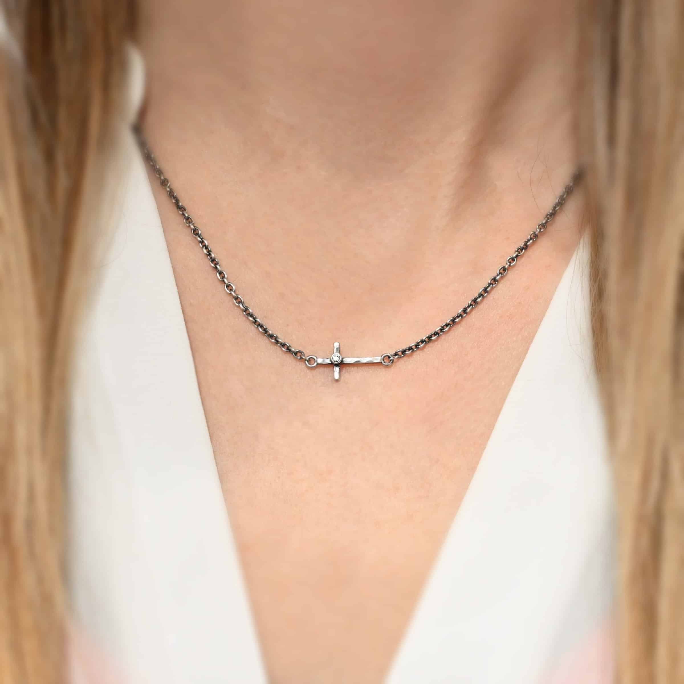 Creator Necklace ox cz Photo of the "Creator" Necklace. Made out of sterling silver. There is a small, but thick hammered silver cross hung vertically and attached on both ends to the chain of the necklace. There is a small cz in the center of the cross. Each end of the cross has a loop on it to attach each end to the chain. Necklace is oxidized and worn up close by a model.