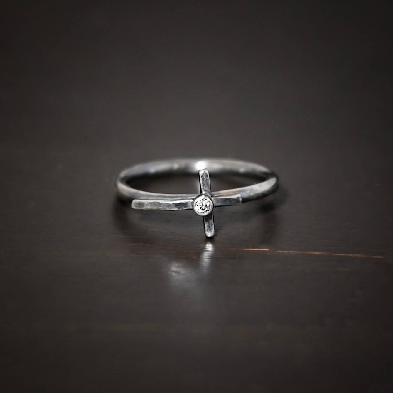 Photo of a creator ring. Ring is made out of sterling silver. The ring band is a thin circle. There is a cross that is in the center of the ring band and set horizontally. The top of bottom of the cross are soldered onto the ring band. The top side of the cross is sticking into the air and the bottom side of the cross is set on the table. There is a small cubic zirconia in the center of the cross. Ring is sitting against a dark background.