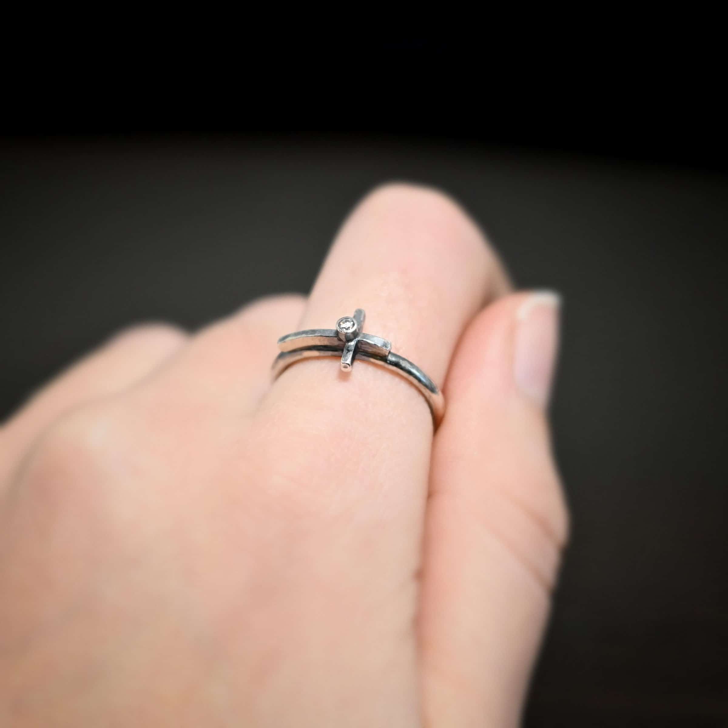 Photo of a creator ring. Ring is made out of sterling silver. The ring band is a thin circle. There is a cross that is in the center of the ring band and set horizontally. The top of bottom of the cross are soldered onto the ring band. The top side of the cross is sticking into the air and the bottom side of the cross is set on the table. There is a small cubic zirconia in the center of the cross. Ring is worn by a model up close.