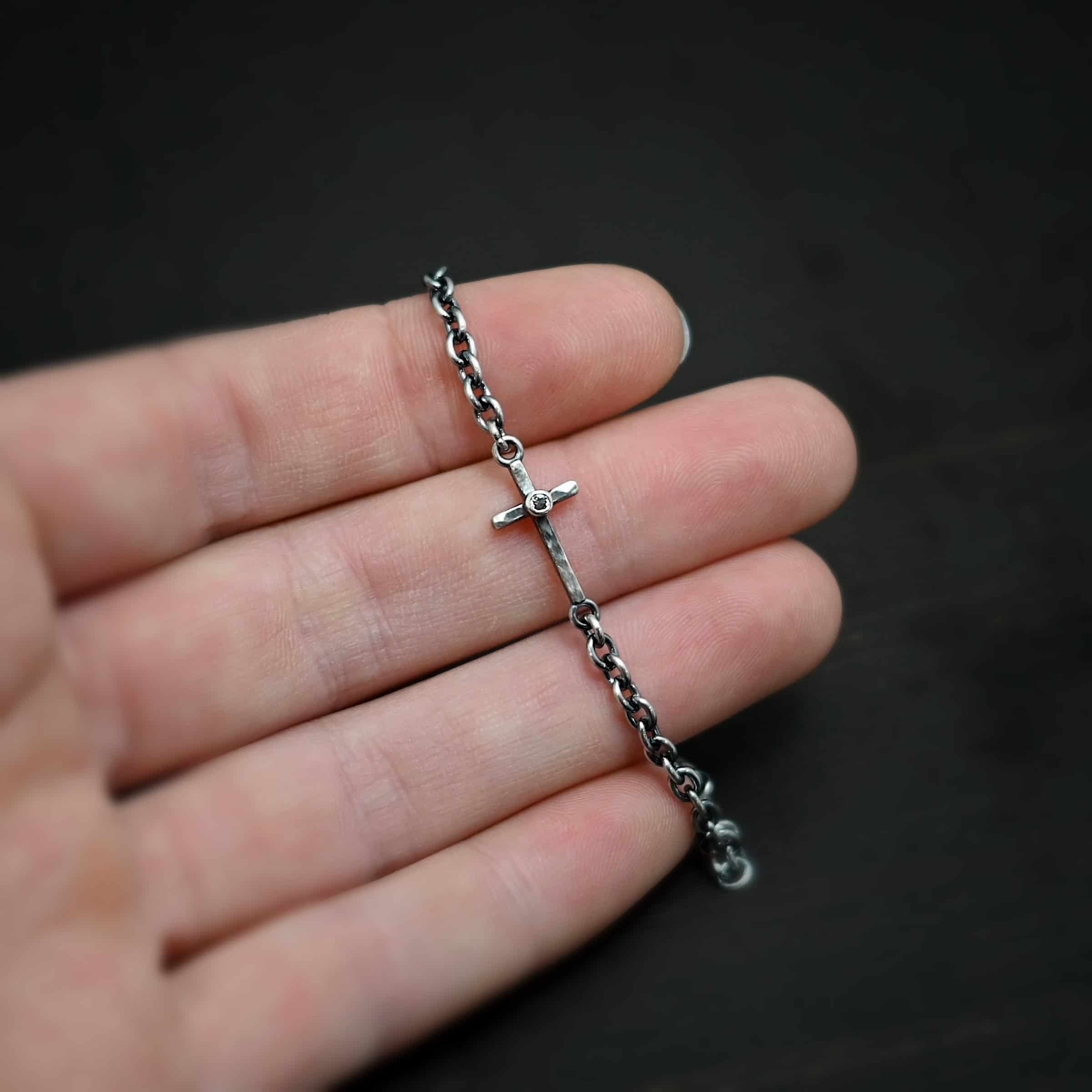 Photo of the "Creator" Bracelet. There is a cross set horizontally with a loop on each end. Each end of the cross is connected to each side of the chain in the center of the bracelet. There is a small cz in the center of the cross. The bracelet is oxidized, and lying against the fingertips of a hand.