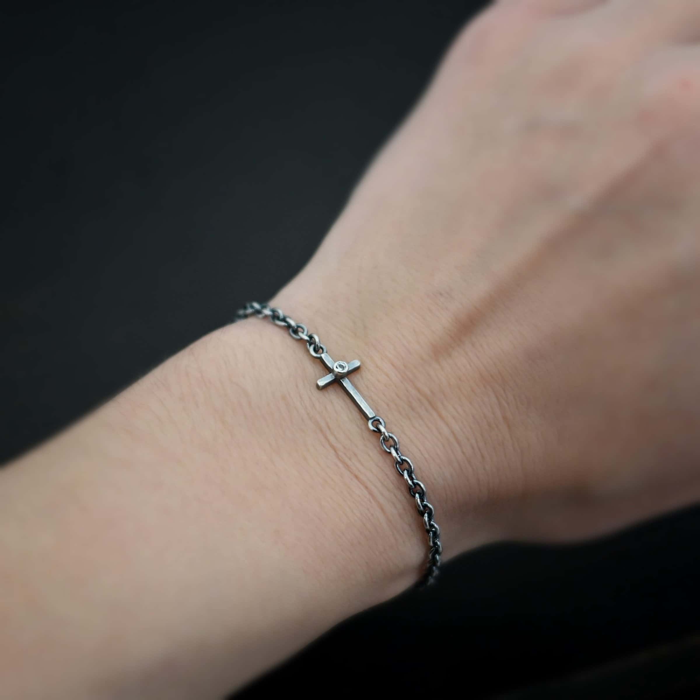 Photo of the "Creator" Bracelet. There is a cross set horizontally with a loop on each end. Each end of the cross is connected to each side of the chain in the center of the bracelet. There is a small cz in the center of the cross. The bracelet is oxidized, and worn up close by a model on their wrist.