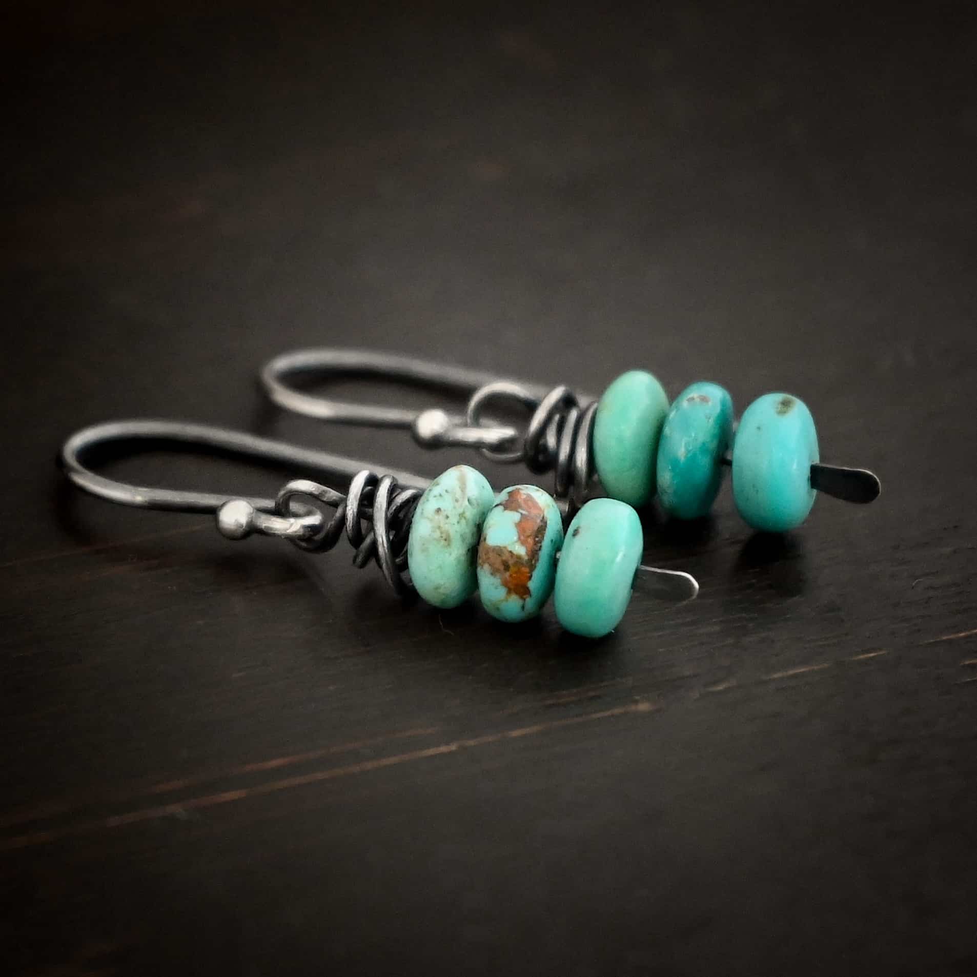 Photo of the "Summer's Breeze" Earrings. Made out of sterling silver and turquoise. Earrings have french hook ear wires. Attached to the ear wires on the bottom is a small silver stick with a loop on the top, silver wrapped around the top of the stick after the loop, and a hammered paddle bottom. In between the wrapped silver and paddle bottom are three small pieces of turquoise. Earrings are lying against a dark background.