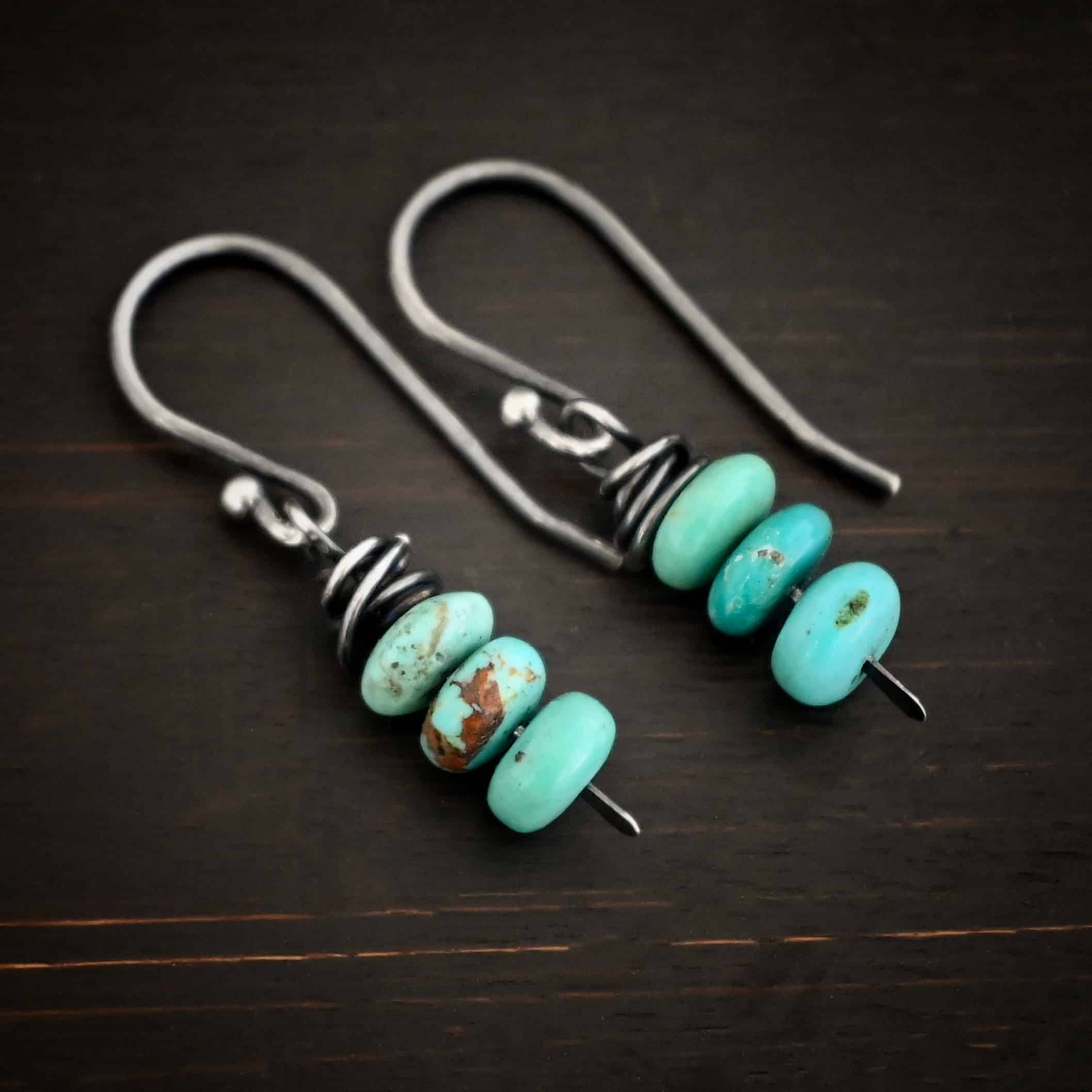 Photo of the "Summer's Breeze" Earrings. Made out of sterling silver and turquoise. Earrings have french hook ear wires. Attached to the ear wires on the bottom is a small silver stick with a loop on the top, silver wrapped around the top of the stick after the loop, and a hammered paddle bottom. In between the wrapped silver and paddle bottom are three small pieces of turquoise. Earrings are lying against a dark background.