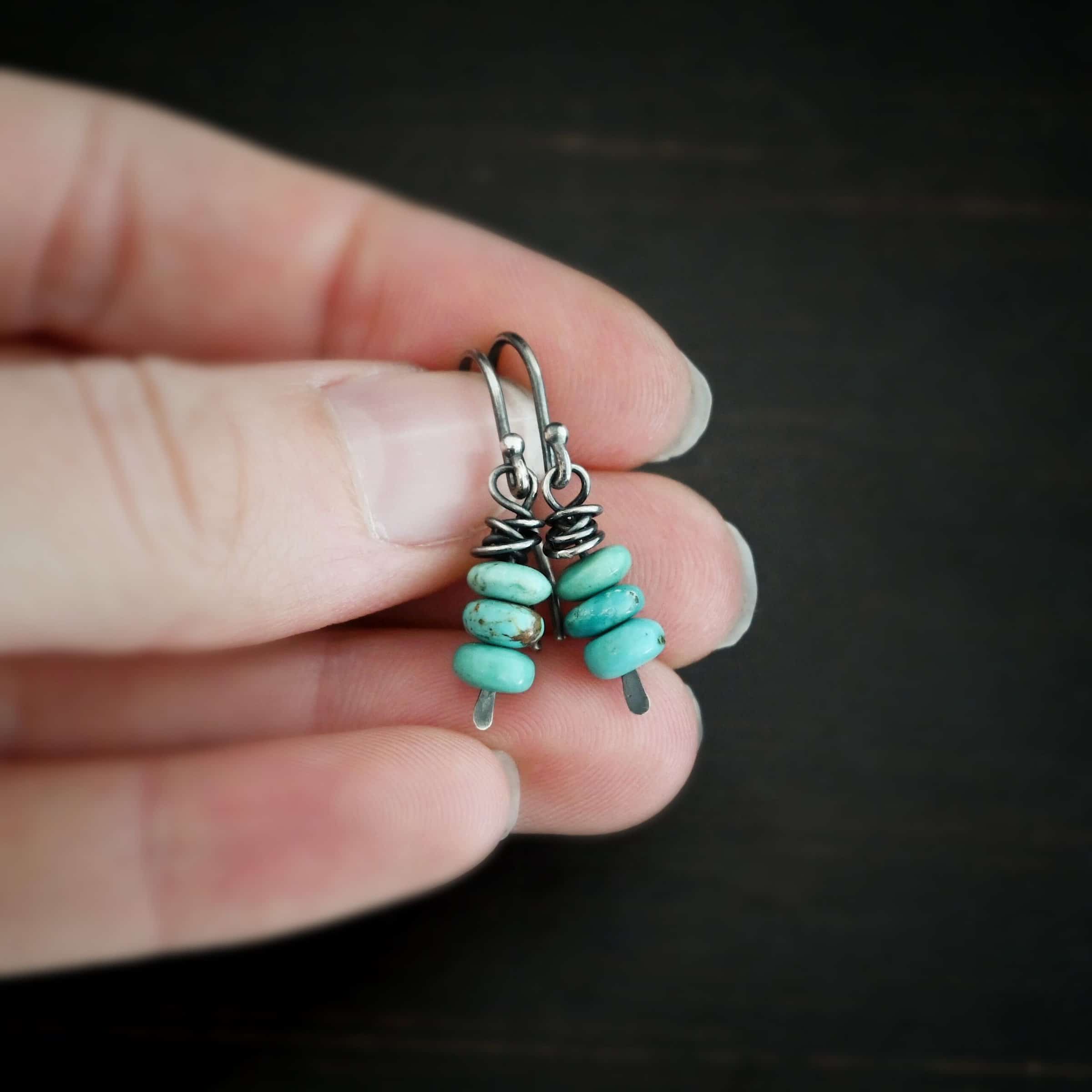 Photo of the "Summer's Breeze" Earrings. Made out of sterling silver and turquoise. Earrings have french hook ear wires. Attached to the ear wires on the bottom is a small silver stick with a loop on the top, silver wrapped around the top of the stick after the loop, and a hammered paddle bottom. In between the wrapped silver and paddle bottom are three small pieces of turquoise. Earrings are held in the fingertips of a hand.