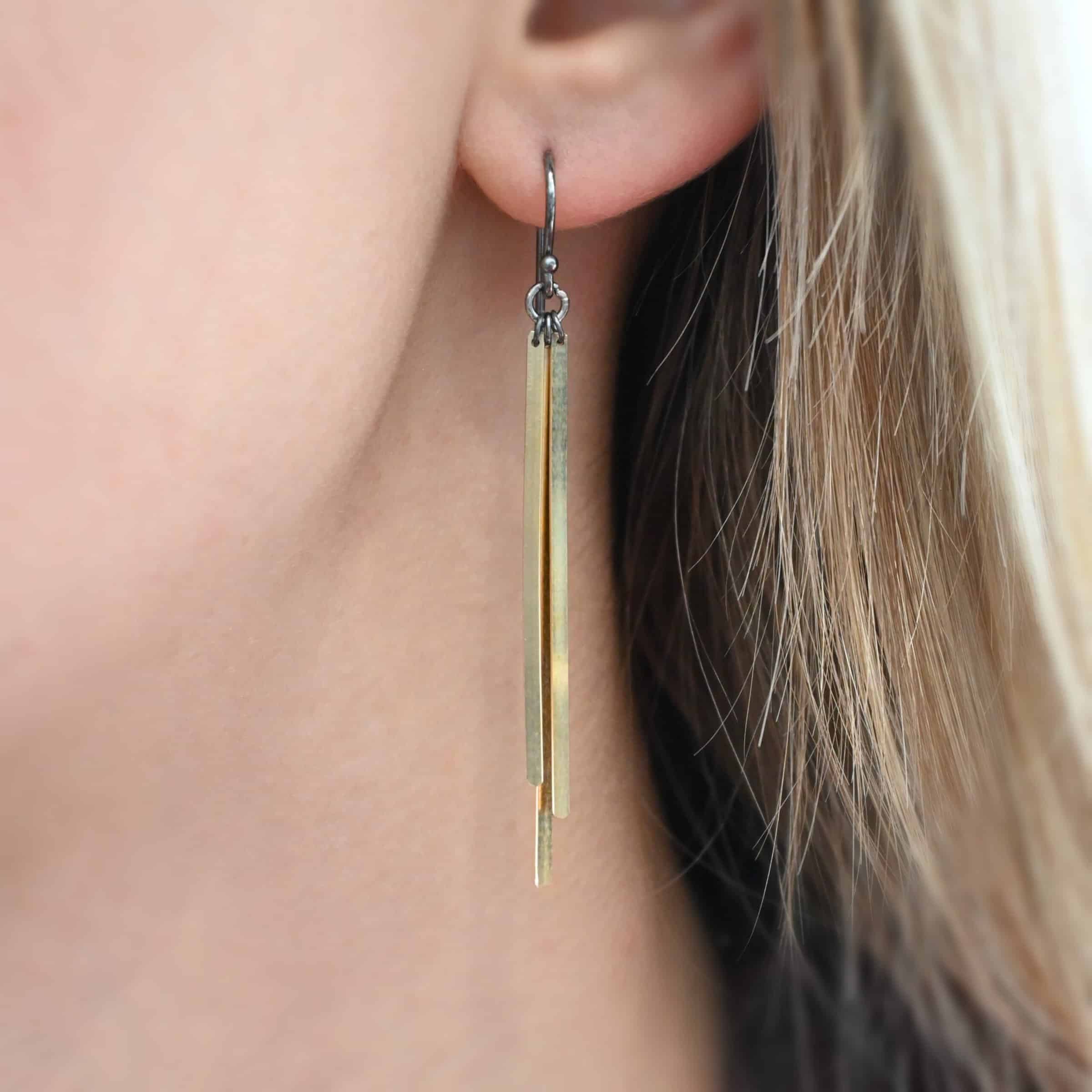 Photo of the "Moonbeam" Earring as shown worn by a model. Made out of sterling silver and 14k gold fill. The earrings are oxidized sterling silver, and have a small silver jump ring attached to the bottom. Attached to the jump ring are three separate pieces of gold fill metal. The pieces are long, flat, thin, rectangles. There are three per earring, each attached to the jump ring on the bottom of the ear wire.