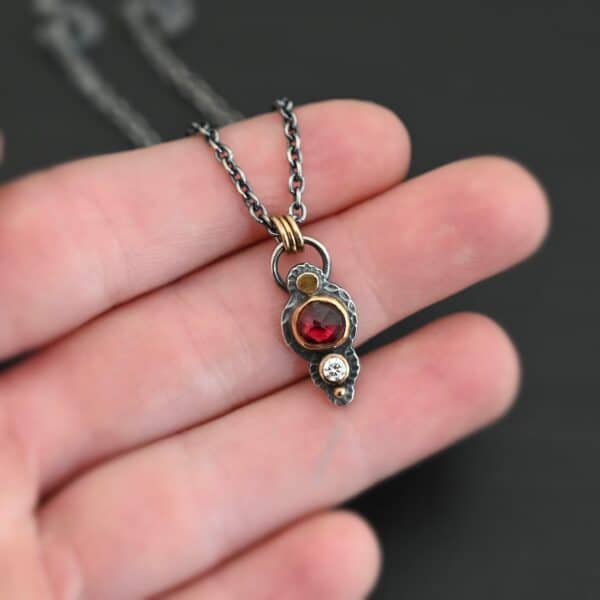 Photo of the "Champagne Blush" Necklace. There is a gold dot at the top, then a small circular garnet set in gold, and then a small diamond that is set in gold, with a gold ball on the very bottom of the pendant. There are three gold jump rings attached to a loop on the top. The chain goes through the jump rings. Necklace is lying against the fingertips of a hand.