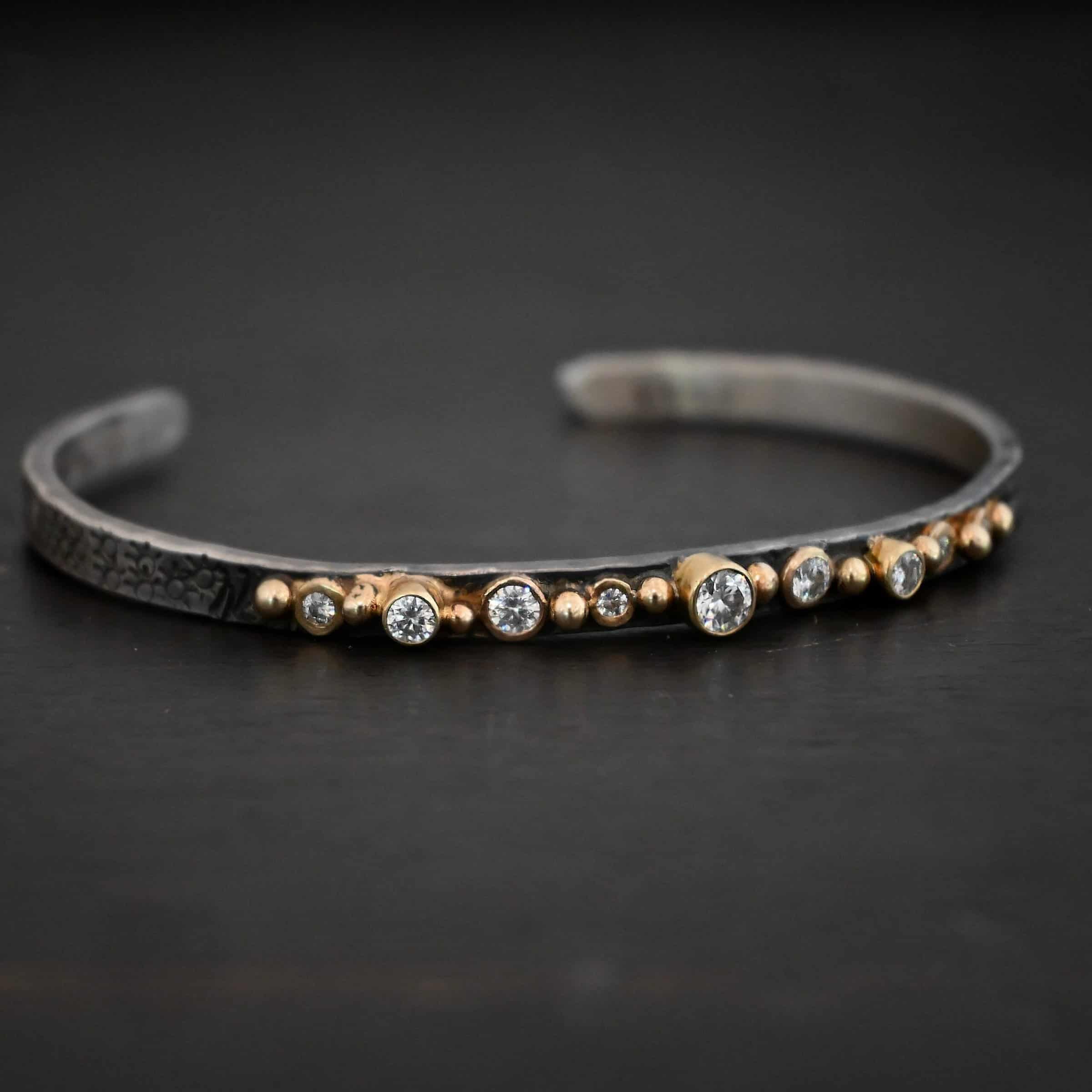 Abrealas Cuff Photo of the "Abrealas" Cuff. Made out of sterling silver, gold, and moissanites. The cuff is silver. There are nine moissanites that go in a line across the cuff. Each one is separated by a gold ball. There are ten gold balls. Cuff is sitting against a dark background.