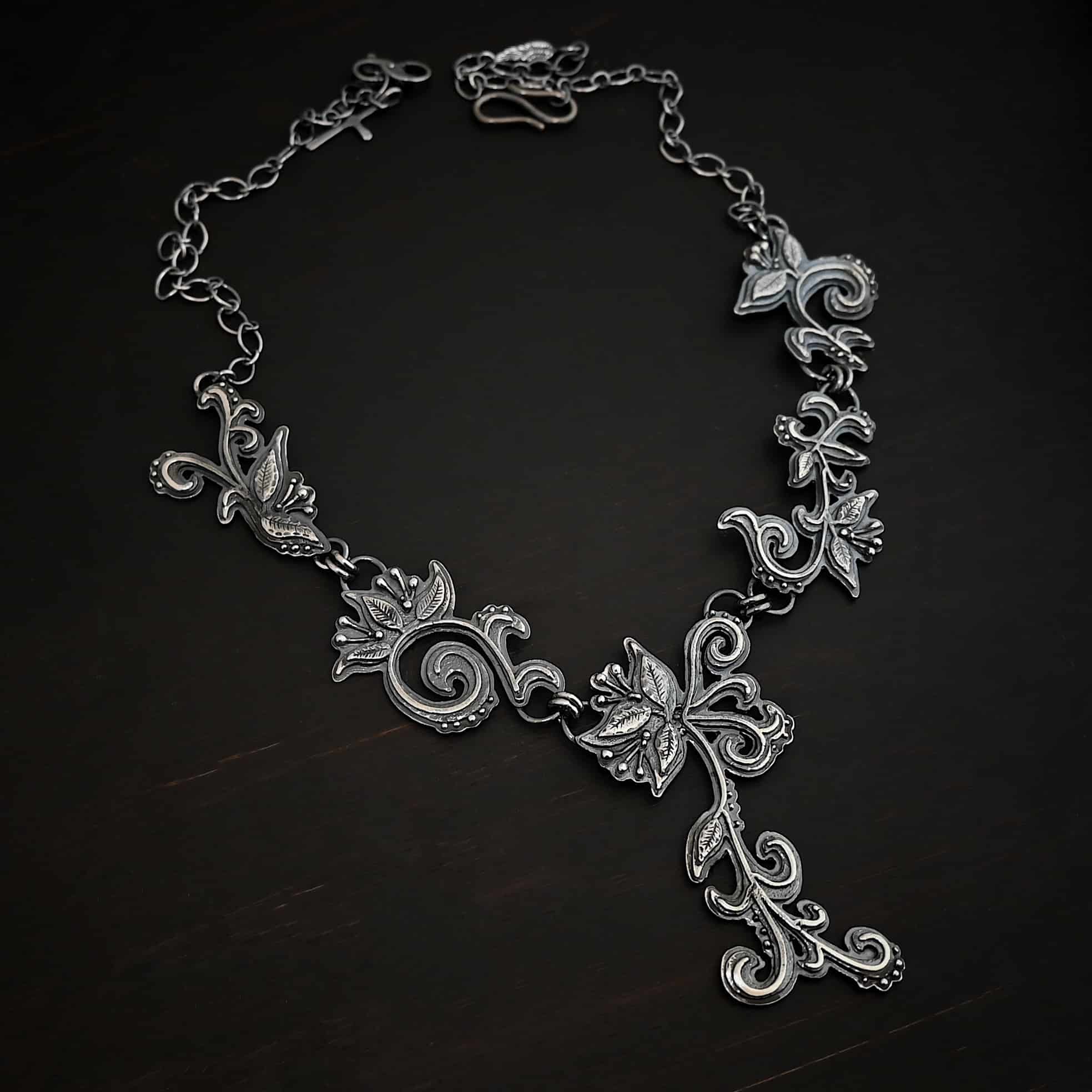 Paisley Necklace Photo of the "Paisley" Necklace. Made out of sterling silver. It is a large collar necklace. The entire necklace is made out of pendants that have sterling silver leaves and silver ball detail around the swirling pendants. The bottom of the necklace has a long, vine like pieces of silver that hangs down, with silver leaves at the top and in the center, and small swirls on the bottom. Necklace is lying against a dark background.