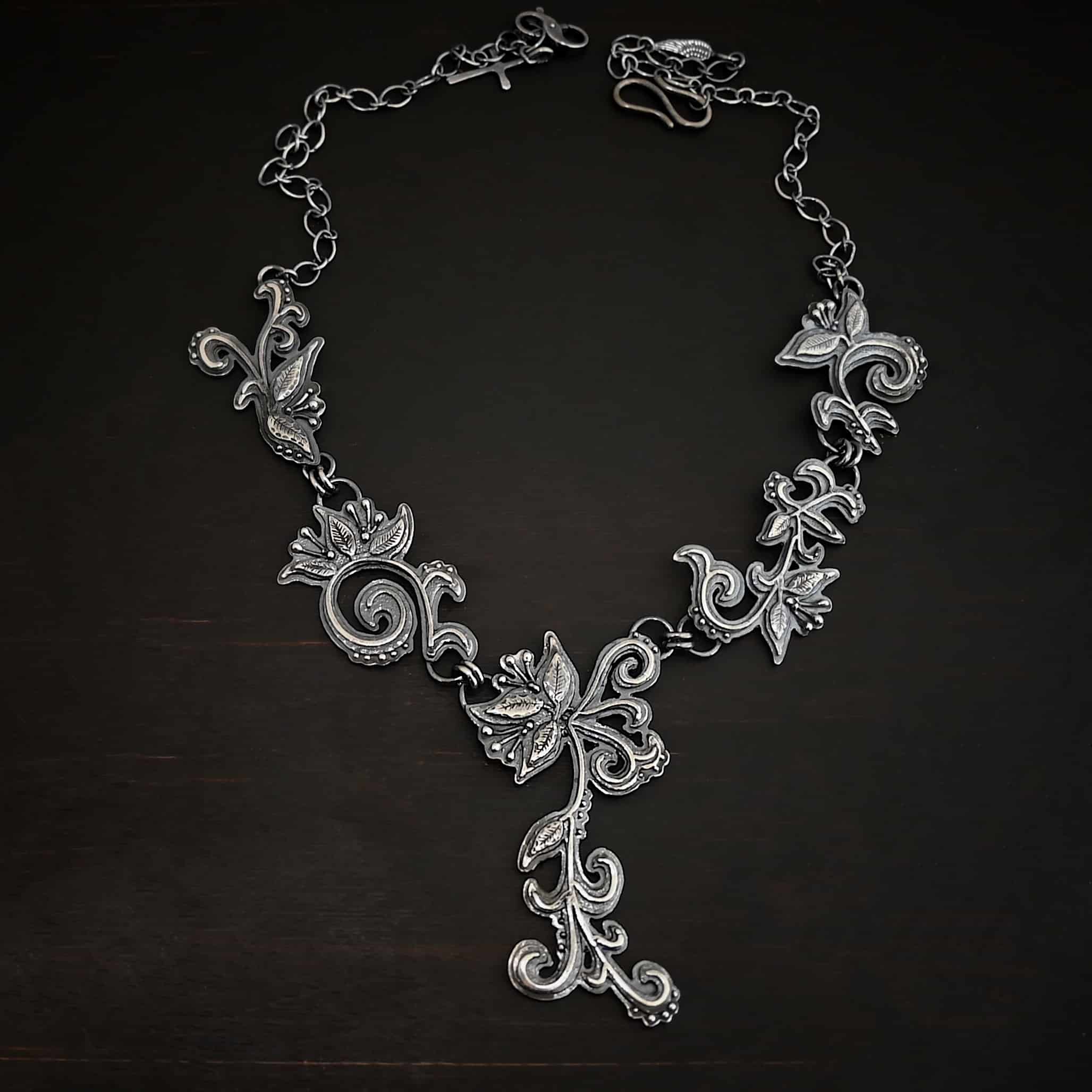 Paisley Necklace Photo of the "Paisley" Necklace. Made out of sterling silver. It is a large collar necklace. The entire necklace is made out of pendants that have sterling silver leaves and silver ball detail around the swirling pendants. The bottom of the necklace has a long, vine like pieces of silver that hangs down, with silver leaves at the top and in the center, and small swirls on the bottom. Necklace is lying against a dark background.