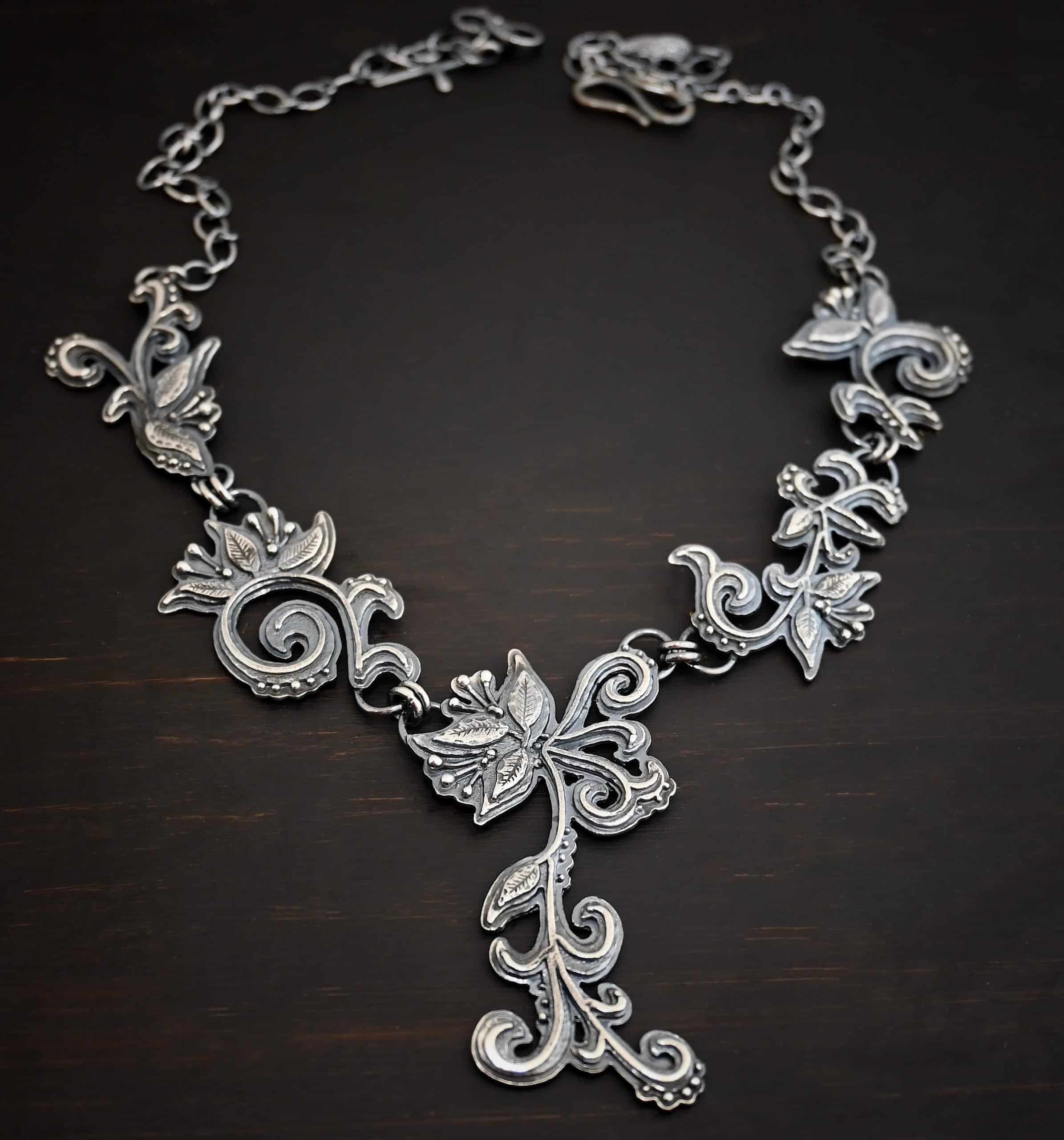 Paisley Necklace Photo of the "Paisley" Necklace. Made out of sterling silver. It is a large collar necklace. The entire necklace is made out of pendants that have sterling silver leaves and silver ball detail around the swirling pendants. The bottom of the necklace has a long, vine like pieces of silver that hangs down, with silver leaves at the top and in the center, and small swirls on the bottom. Necklace is lying against a dark background.
