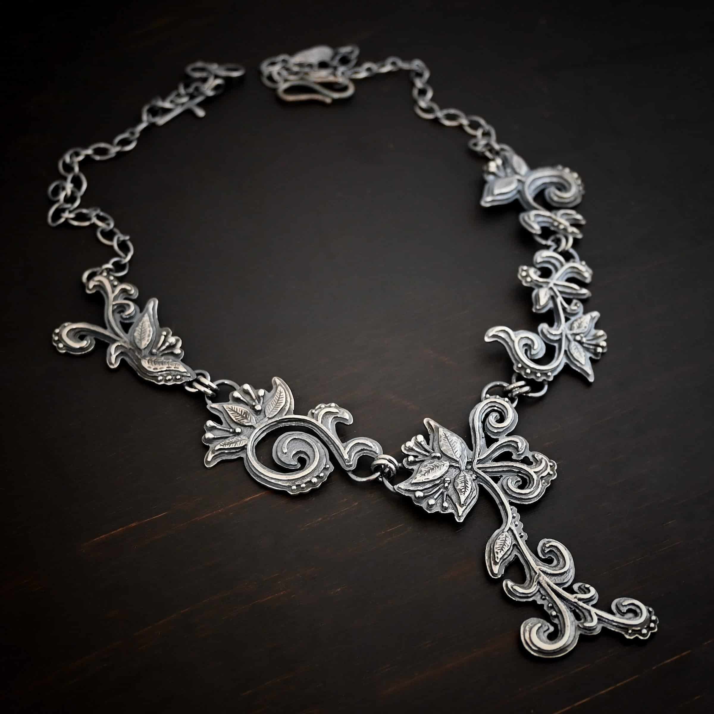 Paisley Necklace Photo of the "Paisley" Necklace. Made out of sterling silver. It is a large collar necklace. The entire necklace is made out of pendants that have sterling silver leaves and silver ball detail around the swirling pendants. The bottom of the necklace has a long, vine like pieces of silver that hangs down, with silver leaves at the top and in the center, and small swirls on the bottom. Necklace is lying against a dark background.