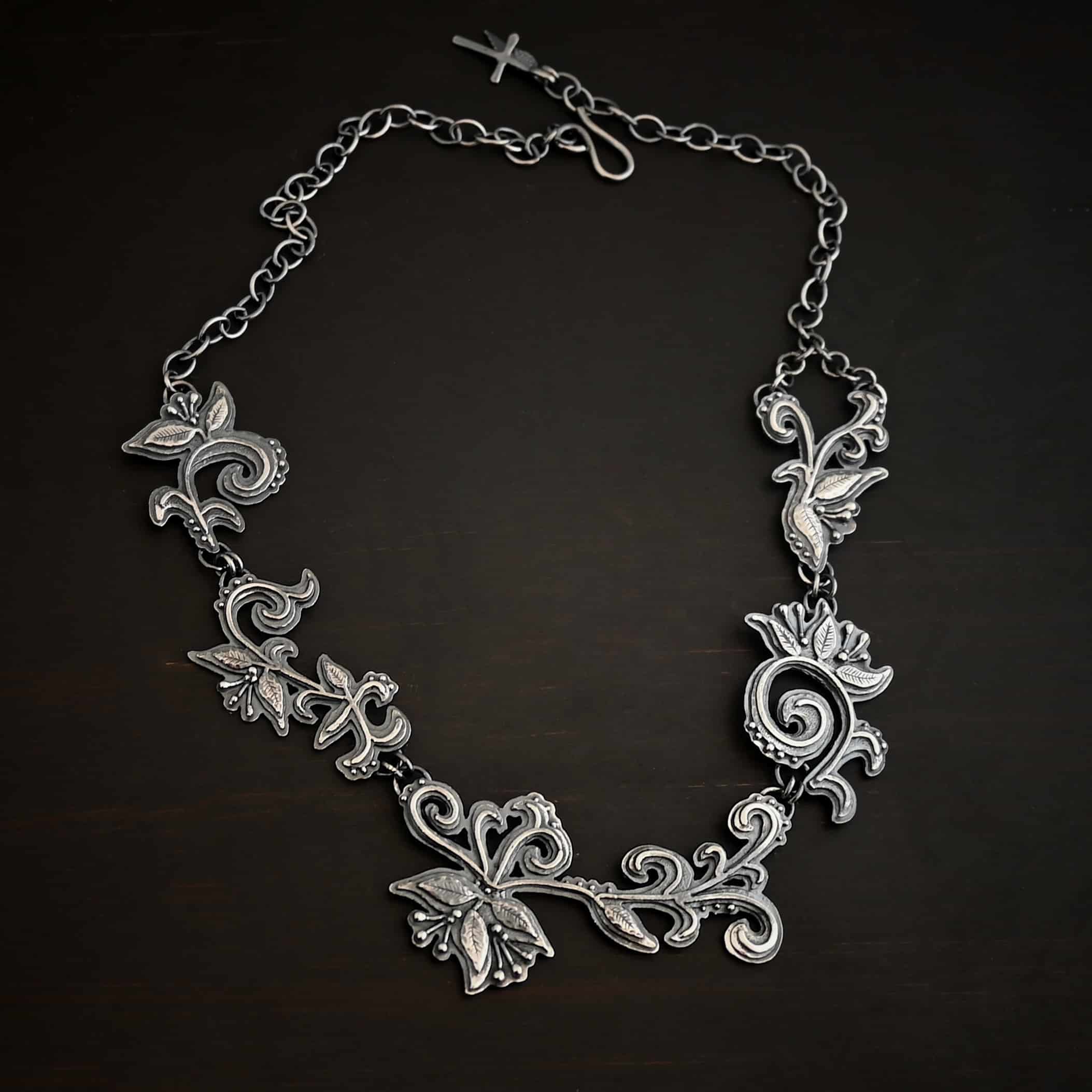 Paisley Necklace Photo of the "Paisley" Necklace. Made out of sterling silver. It is a large collar necklace. The entire necklace is made out of pendants that have sterling silver leaves and silver ball detail around the swirling pendants. The bottom of the necklace has a long, vine like pieces of silver that hangs down, with silver leaves at the top and in the center, and small swirls on the bottom. Necklace is lying against a dark background.