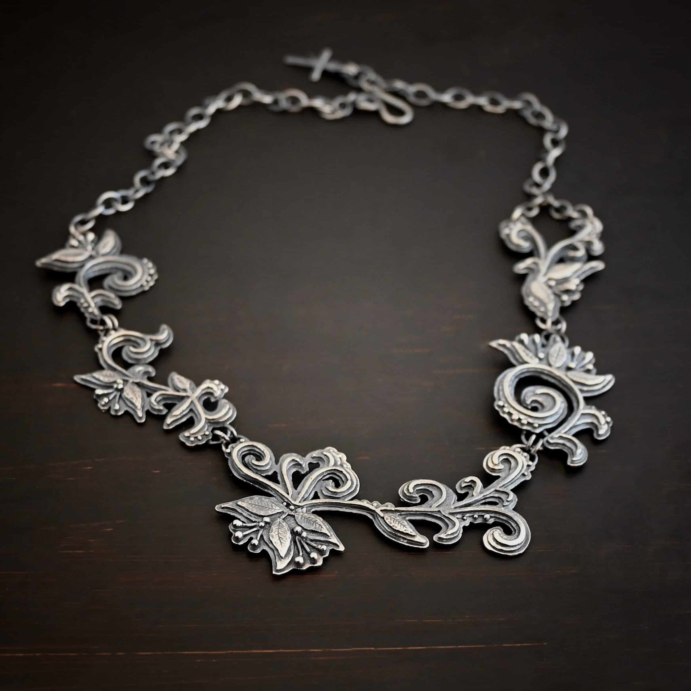 Paisley Necklace Photo of the "Paisley" Necklace. Made out of sterling silver. It is a large collar necklace. The entire necklace is made out of pendants that have sterling silver leaves and silver ball detail around the swirling pendants. The bottom of the necklace has a long, vine like pieces of silver that hangs down, with silver leaves at the top and in the center, and small swirls on the bottom. Necklace is lying against a dark background.