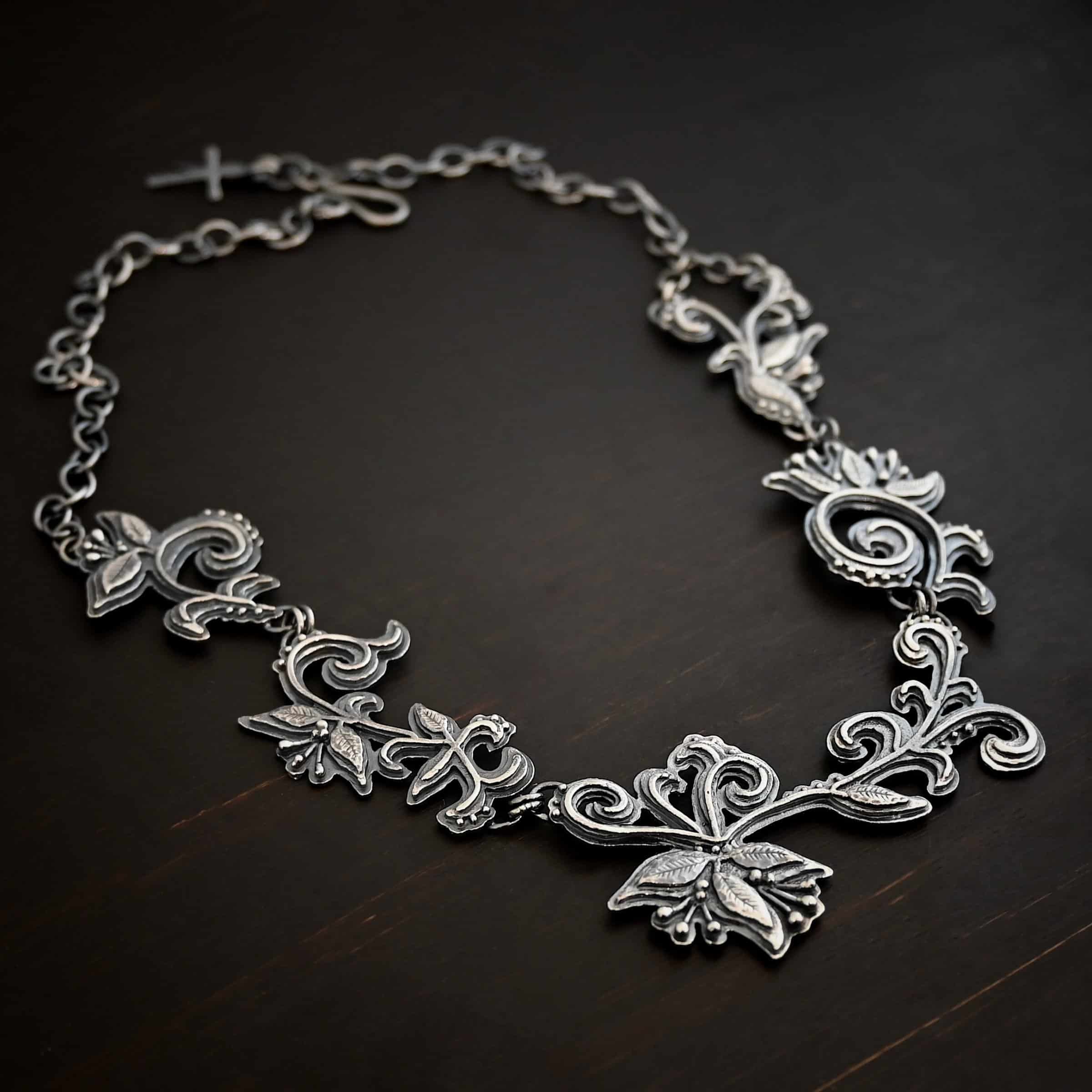 Paisley Necklace Photo of the "Paisley" Necklace. Made out of sterling silver. It is a large collar necklace. The entire necklace is made out of pendants that have sterling silver leaves and silver ball detail around the swirling pendants. The bottom of the necklace has a long, vine like pieces of silver that hangs down, with silver leaves at the top and in the center, and small swirls on the bottom. Necklace is lying against a dark background.