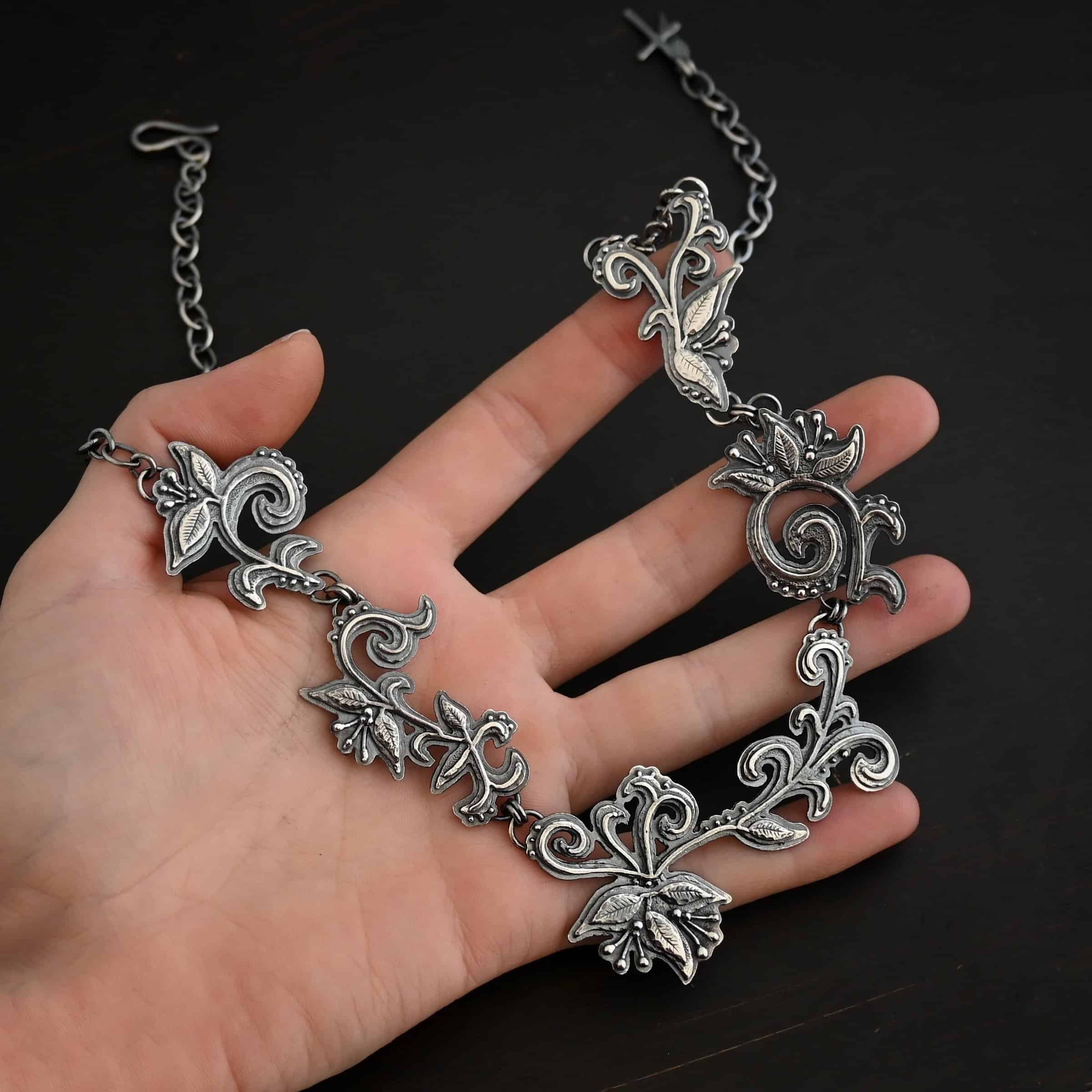 Paisley Necklace Photo of the "Paisley" Necklace. Made out of sterling silver. It is a large collar necklace. The entire necklace is made out of pendants that have sterling silver leaves and silver ball detail around the swirling pendants. The bottom of the necklace has a long, vine like pieces of silver that hangs down, with silver leaves at the top and in the center, and small swirls on the bottom. Necklace is lying against the fingertips of a hand.