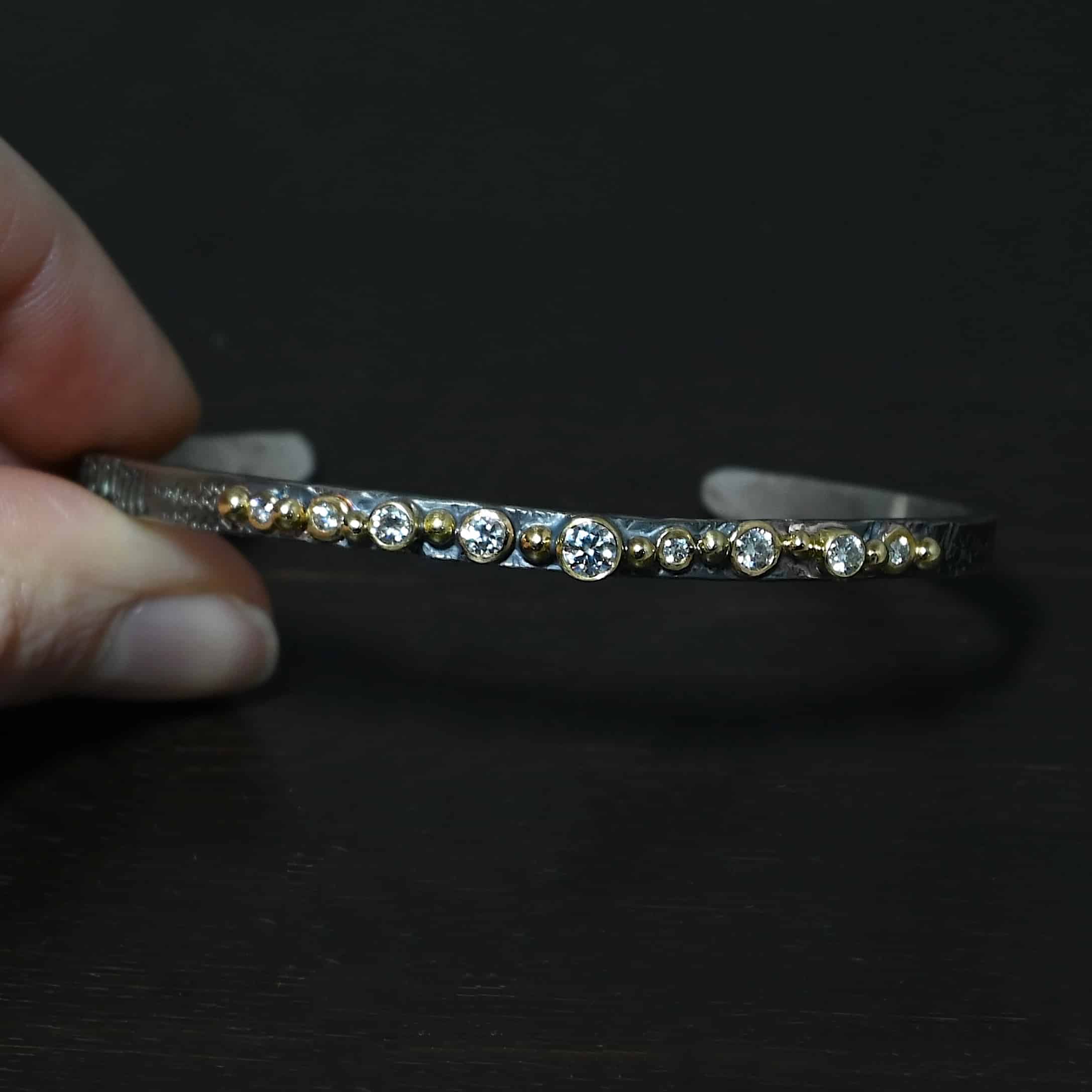 Abrealas Cuff Photo of the "Abrealas" Cuff. Made out of sterling silver, gold, and moissanites. The cuff is silver. There are nine moissanites that go in a line across the cuff. Each one is separated by a gold ball. There are ten gold balls. Cuff is sitting against a dark background and is held in the fingertips of a hand.