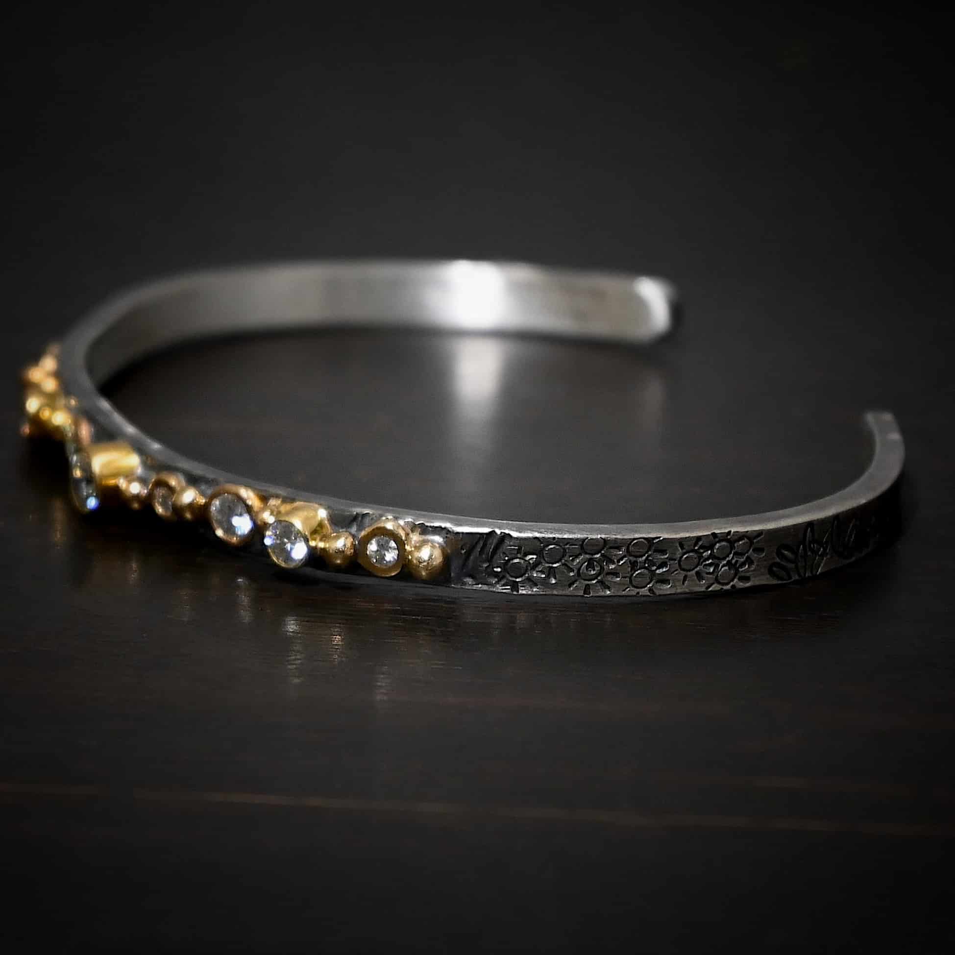 Abrealas Cuff Photo of the "Abrealas" Cuff. Made out of sterling silver, gold, and moissanites. The cuff is silver. There are nine moissanites that go in a line across the cuff. Each one is separated by a gold ball. There are ten gold balls. Cuff is sitting against a dark background.