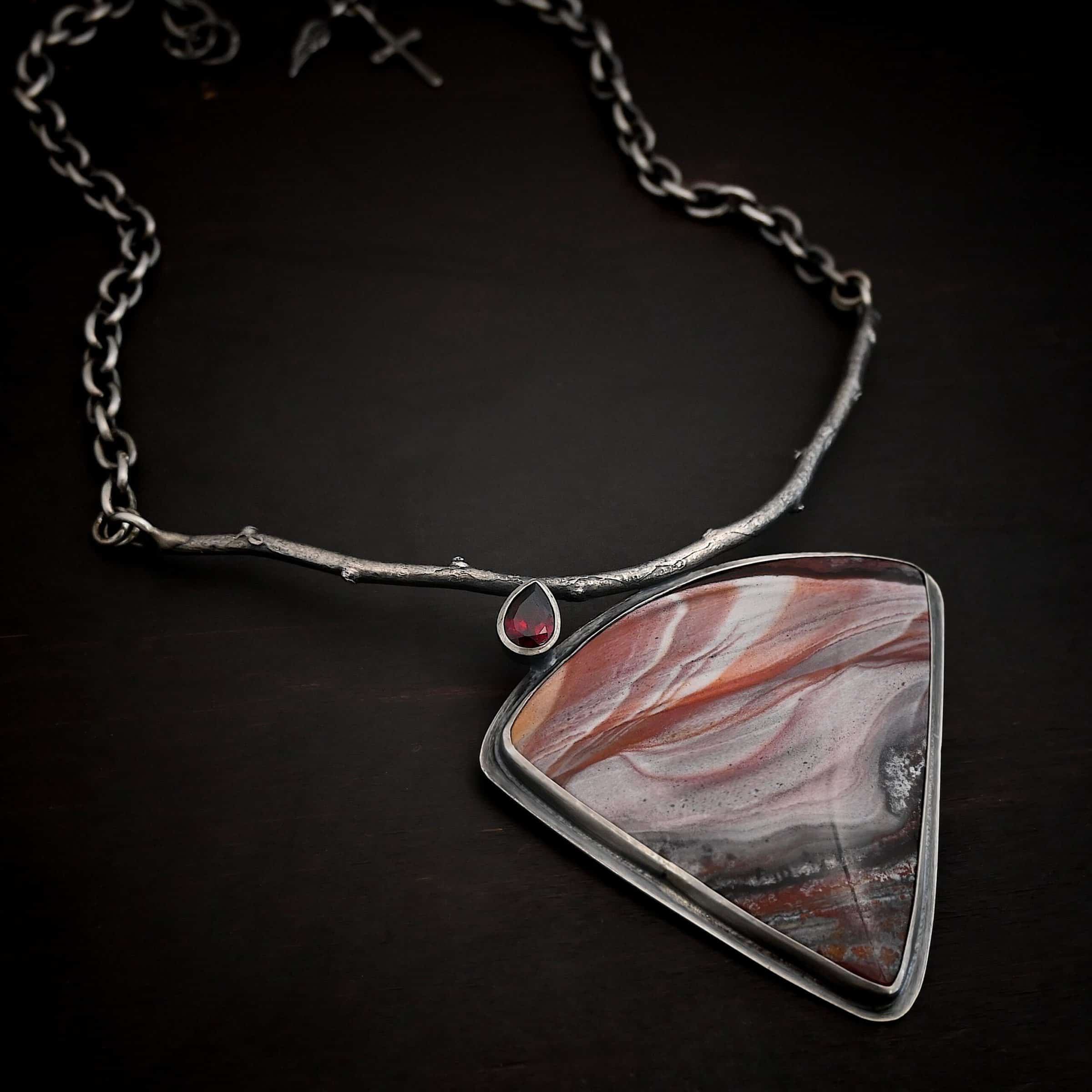 Borrowed from someone beautiful necklace Close up of the "Borrowed From Someone Beautiful" Necklace. Made out of sterling silver, landscape jasper, and garnet. The landscape jasper is the largest stone. It is a triangle shape and is a mix of red, and gray colors, with touches of orange, pink, and white. There is a teardrop shaped garnet on top of the jasper on the left side of the pendant. The point of the teardrop is attached to a casted sterling silver "branch." The bottom of the teardrop is attached to the jasper. Pendant is laying against a dark background.