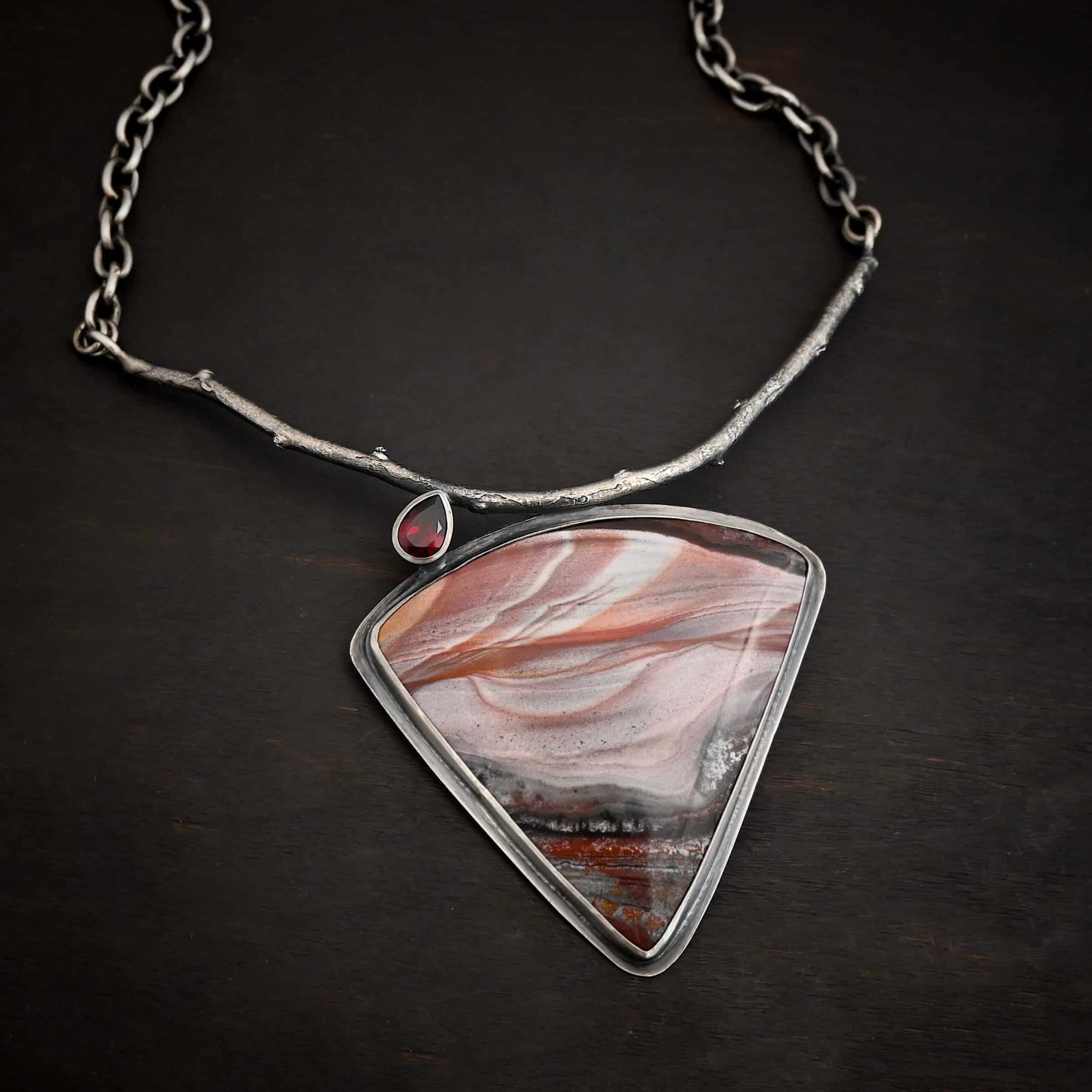 Borrowed from someone beautiful necklace Close up of the "Borrowed From Someone Beautiful" Necklace. Made out of sterling silver, landscape jasper, and garnet. The landscape jasper is the largest stone. It is a triangle shape and is a mix of red, and gray colors, with touches of orange, pink, and white. There is a teardrop shaped garnet on top of the jasper on the left side of the pendant. The point of the teardrop is attached to a casted sterling silver "branch." The bottom of the teardrop is attached to the jasper. Pendant is laying against a dark background.