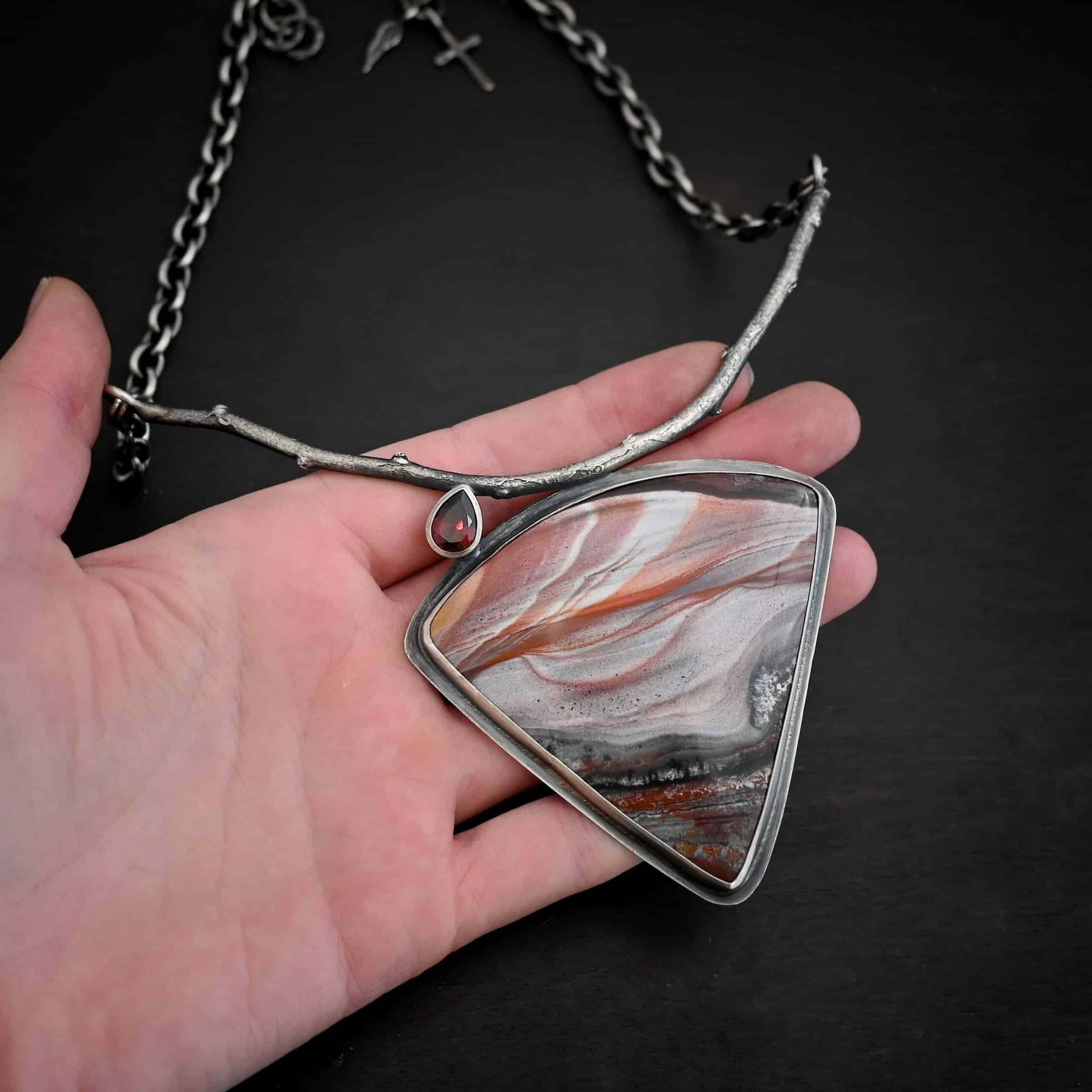 Borrowed from someone beautiful necklace Close up of the "Borrowed From Someone Beautiful" Necklace. Made out of sterling silver, landscape jasper, and garnet. The landscape jasper is the largest stone. It is a triangle shape and is a mix of red, and gray colors, with touches of orange, pink, and white. There is a teardrop shaped garnet on top of the jasper on the left side of the pendant. The point of the teardrop is attached to a casted sterling silver "branch." The bottom of the teardrop is attached to the jasper. Pendant is laying against the fingertips of a hand.