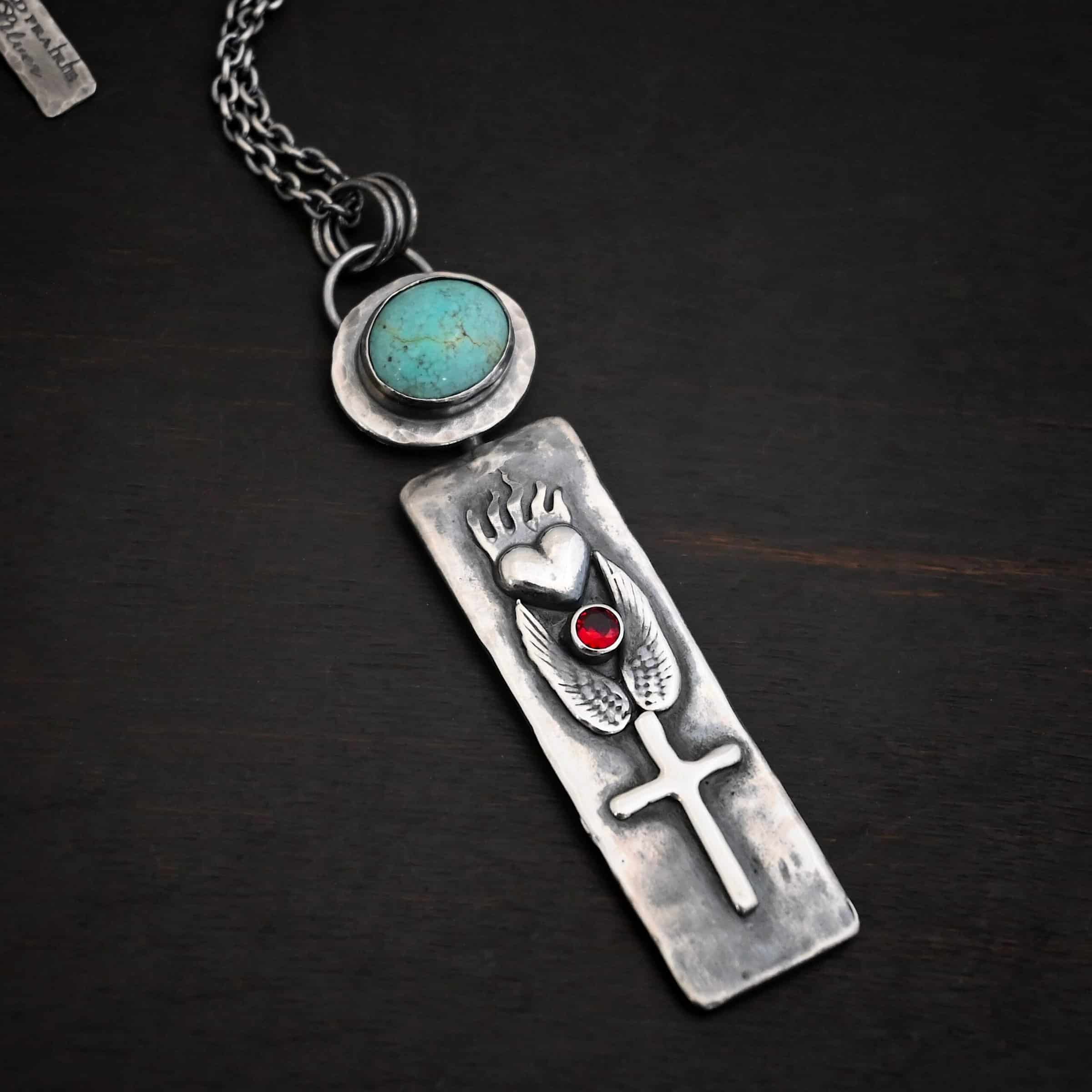 Photo of the "Sacred Heart" Necklace. Necklace is made out of silver, garnet, and turquoise. The pendant is a rectangle shaped silver tag that hangs vertically. It is large. On the top of the rectangle is a silver heart with flames dancing across the top of it. Beneath the heart on the bottom tip of it is a circular small garnet. Beneath this are two silver angel wings, one on each side of the garnet surrounding it. Beneath the angel wings in the center of them and on the bottom of the rectangle pendant is a silver cross soldered to the pendant. There is an oval shaped turquoise soldered to the top of the rectangular pendant. The silver around the turquoise is hammered and oxidized. There is a loop on the back of the turquoise with three jump rings going through it. The chain of the necklace goes through the jump rings. Necklace is lying against a dark background.