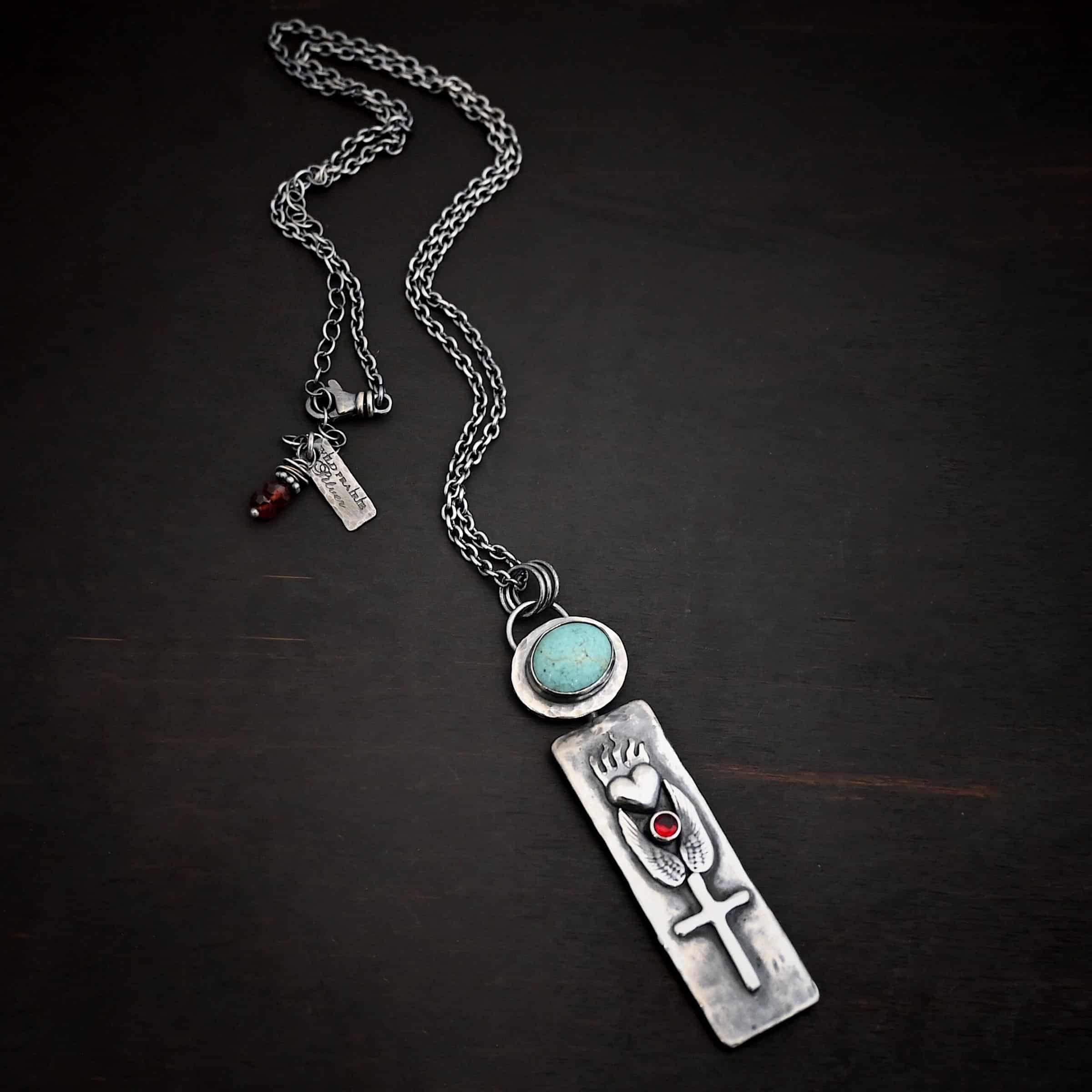 Photo of the "Sacred Heart" Necklace. Necklace is made out of silver, garnet, and turquoise. The pendant is a rectangle shaped silver tag that hangs vertically. It is large. On the top of the rectangle is a silver heart with flames dancing across the top of it. Beneath the heart on the bottom tip of it is a circular small garnet. Beneath this are two silver angel wings, one on each side of the garnet surrounding it. Beneath the angel wings in the center of them and on the bottom of the rectangle pendant is a silver cross soldered to the pendant. There is an oval shaped turquoise soldered to the top of the rectangular pendant. The silver around the turquoise is hammered and oxidized. There is a loop on the back of the turquoise with three jump rings going through it. The chain of the necklace goes through the jump rings. Necklace is lying against a dark background.